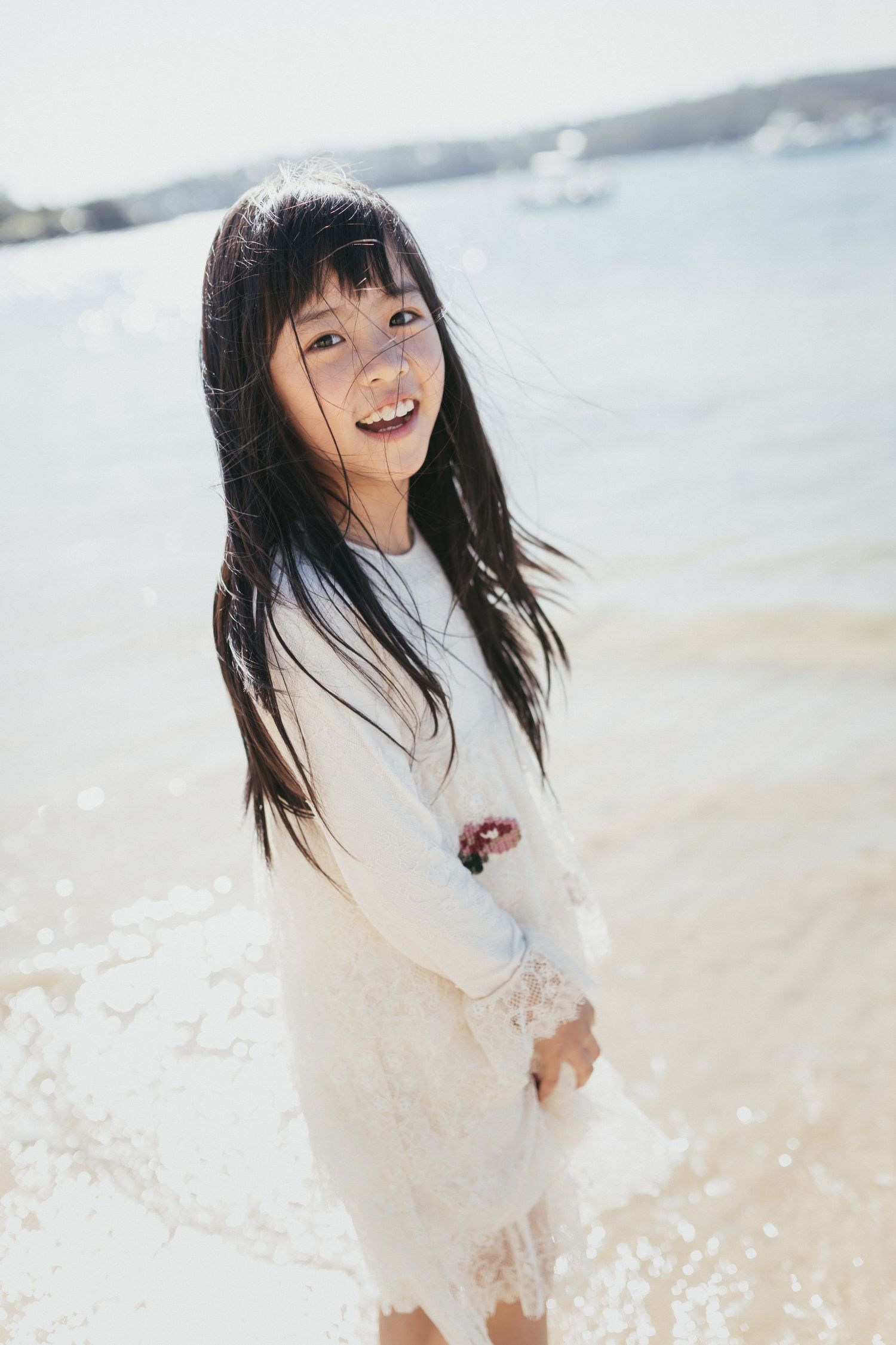 Smiling young Asian girl with long dark hair standing in shallow ocean water wearing a white dress.