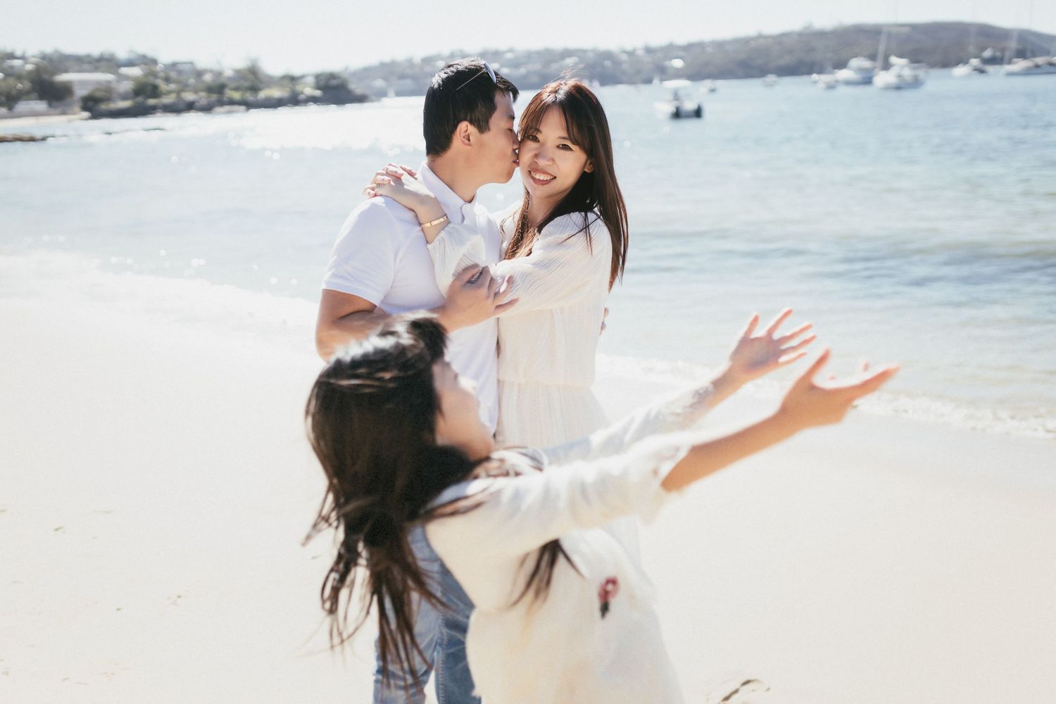 Happy Asian family on a sunny beach, parents embracing while young daughter runs toward them.