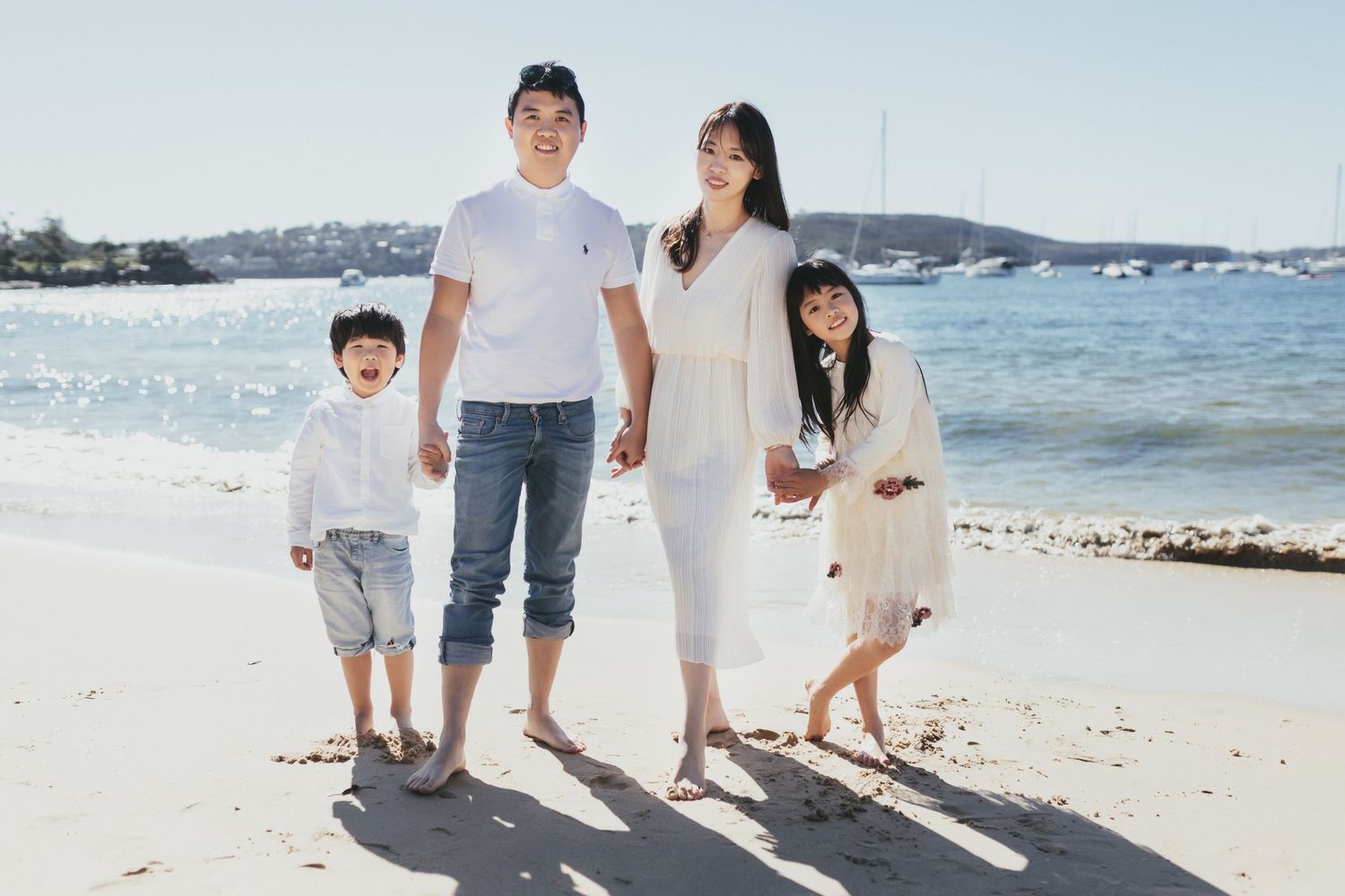 A happy Asian family of four poses together on a sunny beach with calm blue water behind them.