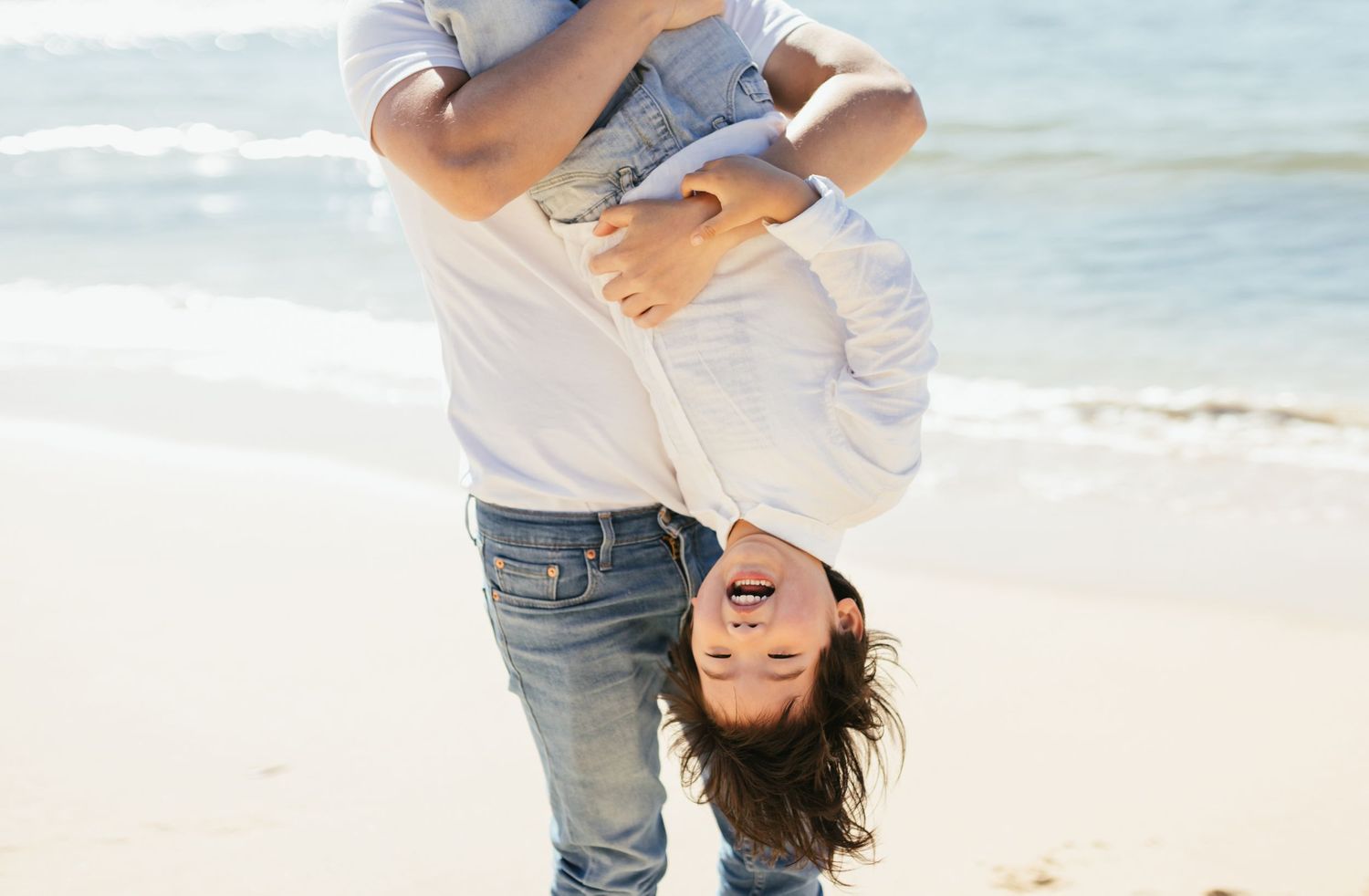 A parent playfully holds a child upside down on a sunny beach with ocean waves in the background.