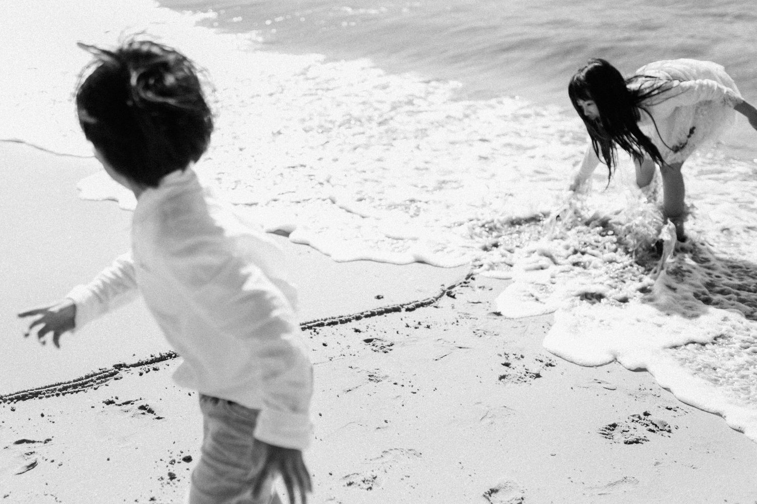 Two children play in black and white photo at the ocean's edge, splashing in the waves on a beach.