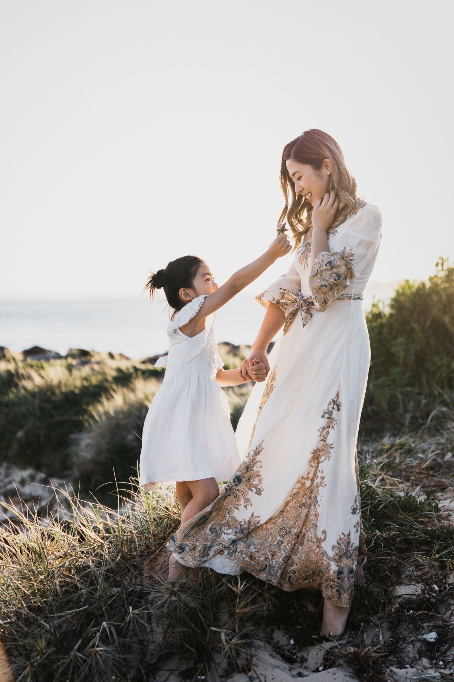 A mother in a floral gown dances with her young daughter in a white dress at a coastal sunset.