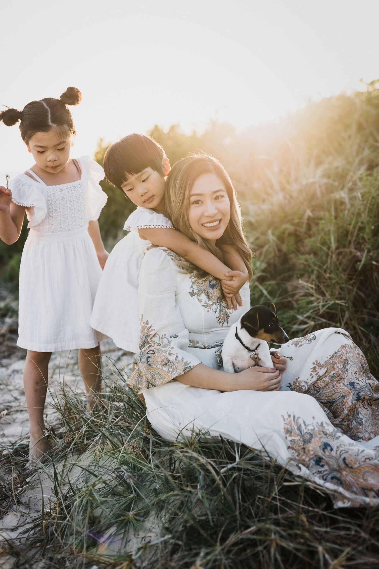 A smiling Asian mother sits in beach grass at sunset with her two children and a small dog.