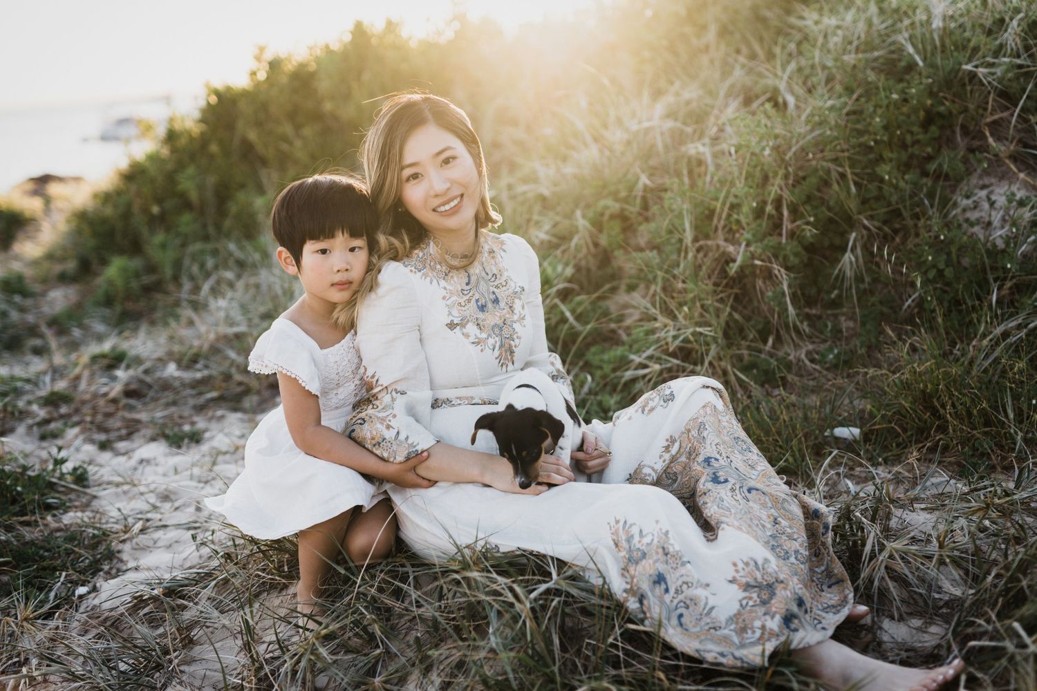 A mother and young daughter sit together in golden beach grass holding a small puppy at sunset.