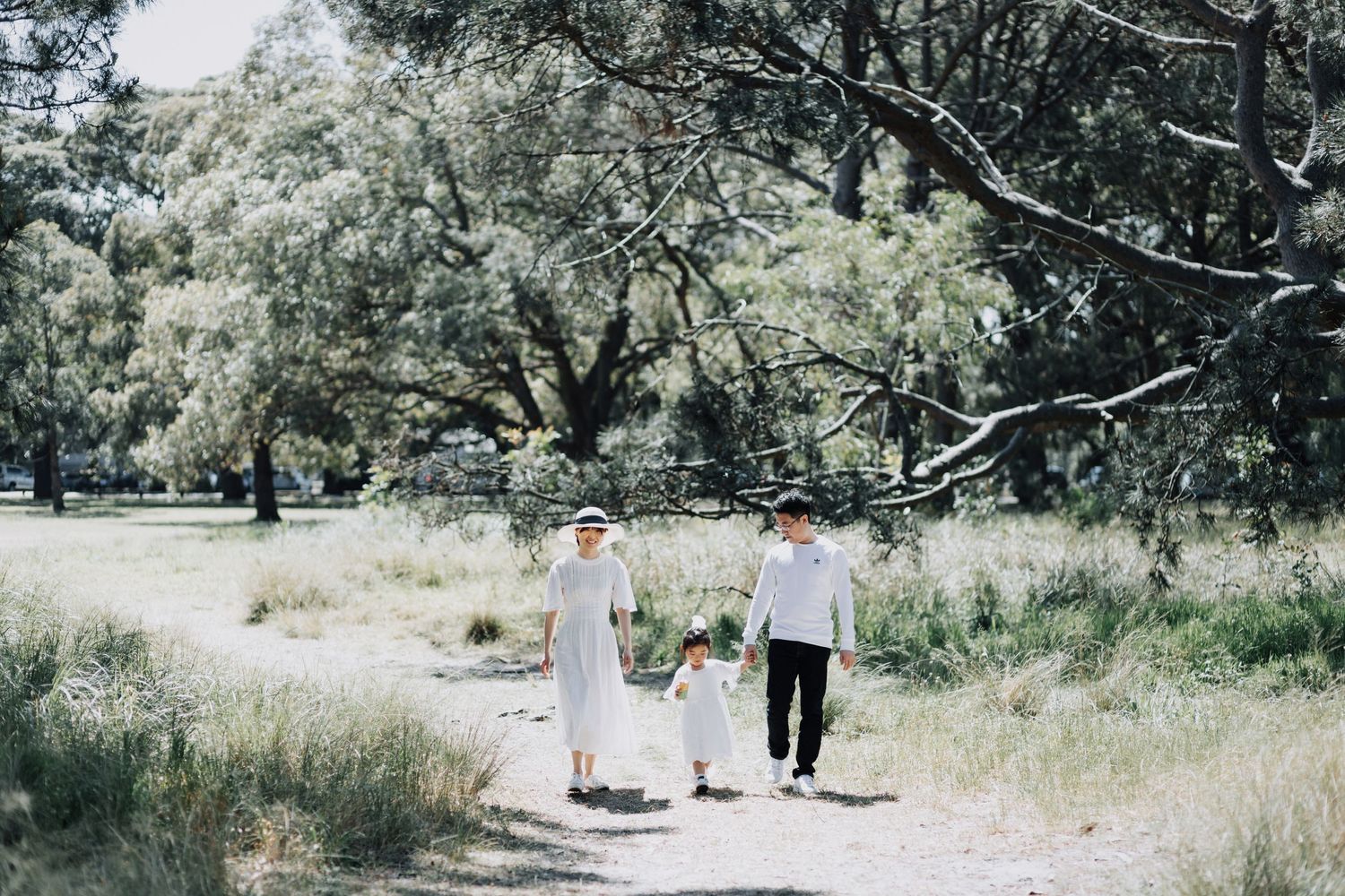 A family of three walks hand-in-hand along a sunlit path through a lush green parkland setting.