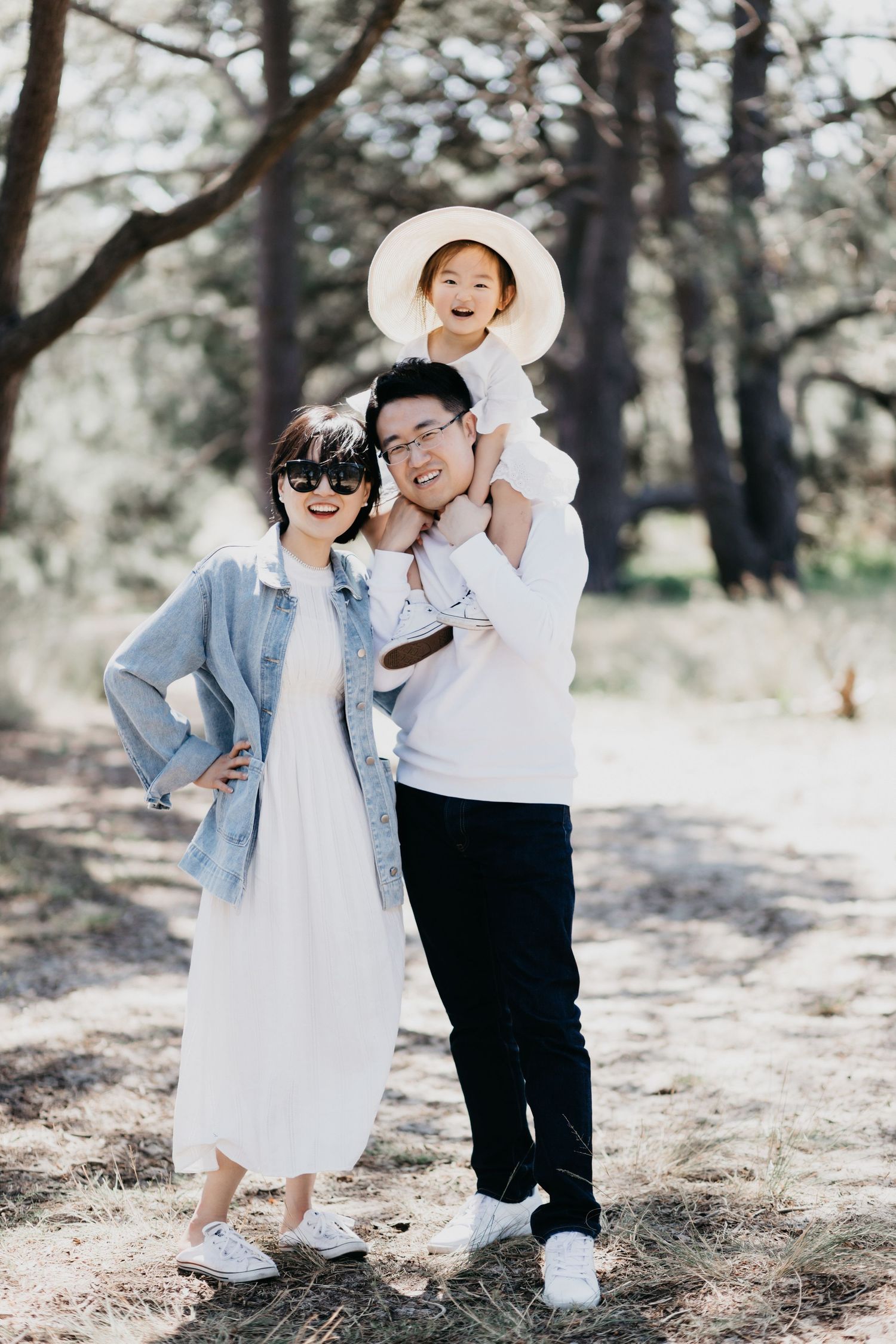 A smiling Asian family poses in a sunlit forest, with their child sitting on the father's shoulders.