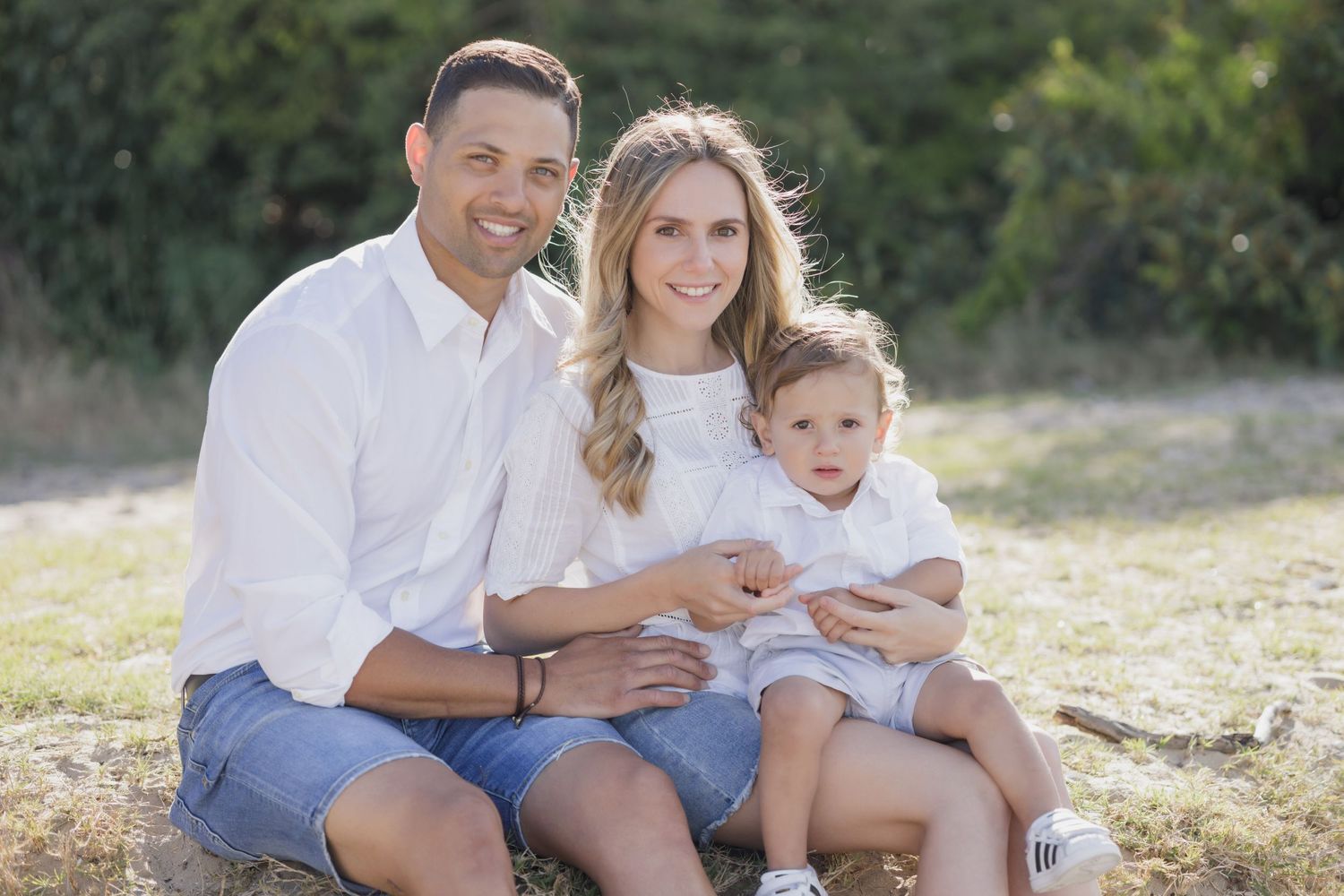 Happy family of three sitting on grass outdoors, parents in white shirts with young child between them.