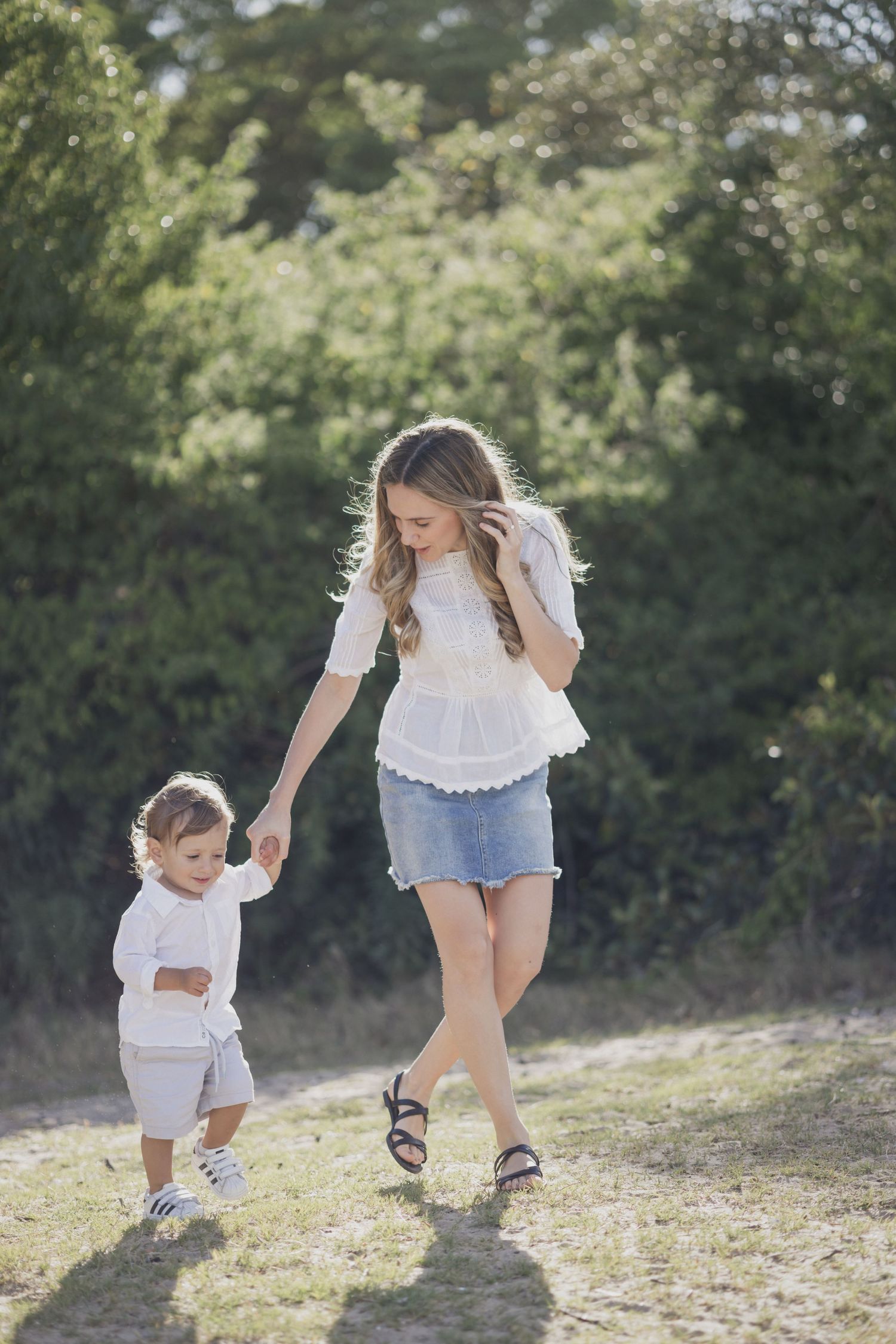 Young mother in white top and denim skirt walking outdoors with toddler on a sunny day.
