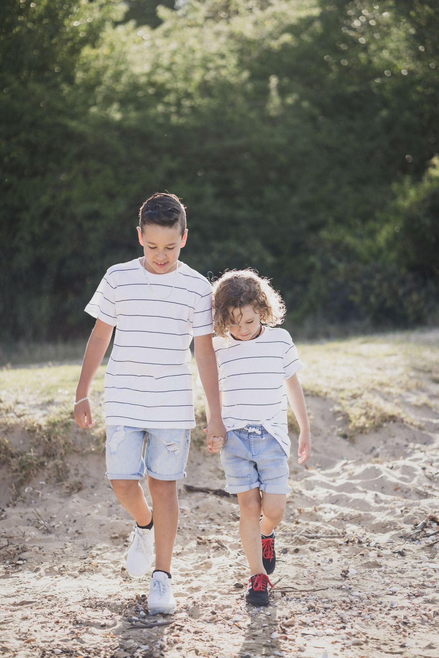Two brothers in matching striped shirts and denim shorts walking hand in hand on a sandy path.