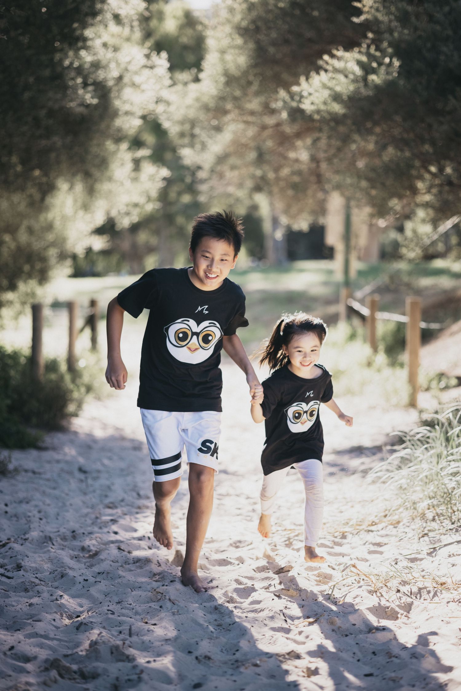 Two Asian children in matching black owl shirts run barefoot along a sandy tree-lined beach path.