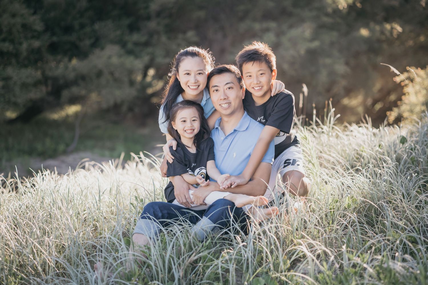 A happy Asian family of four poses together in a sunlit grassy field for a professional portrait.