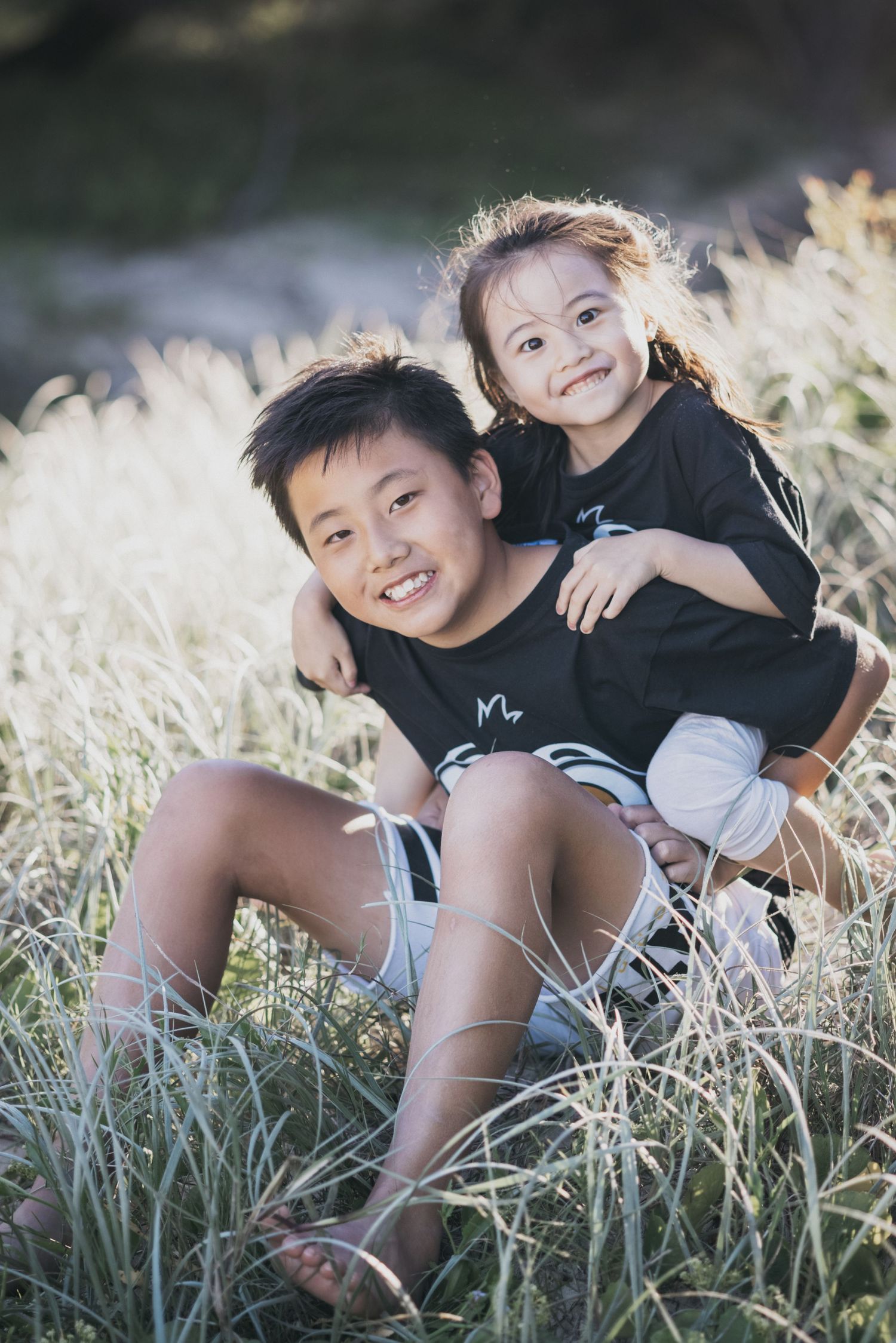 A smiling Asian boy and little girl in matching black outfits sit together in a sunlit grassy field.