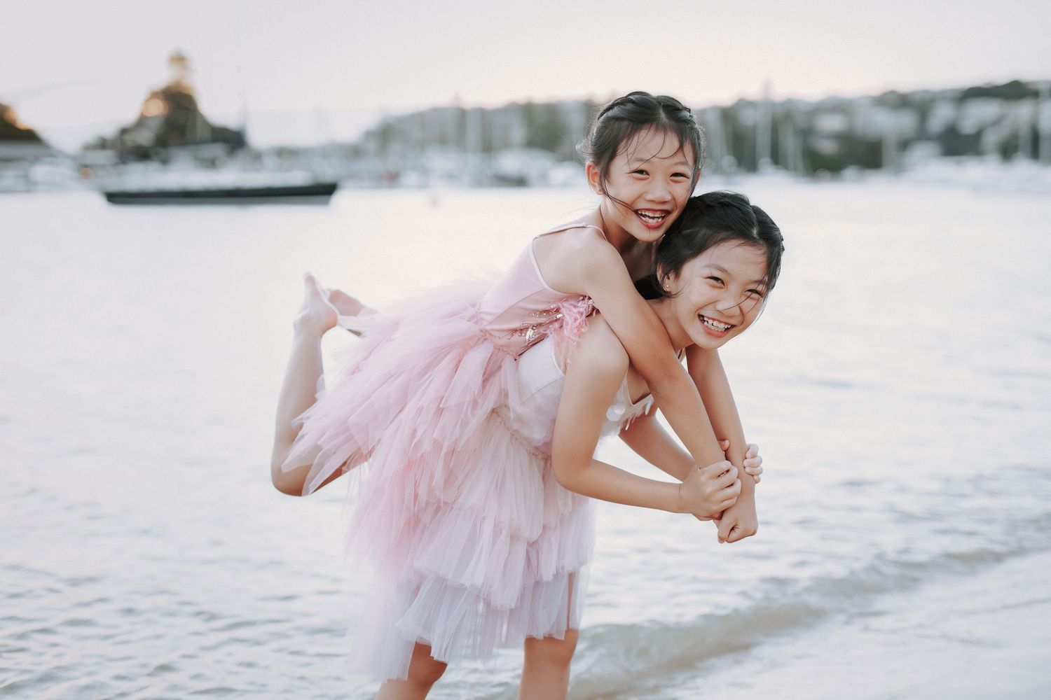 Two young Asian girls in pink dresses laugh as one gives the other a piggyback ride by the water.