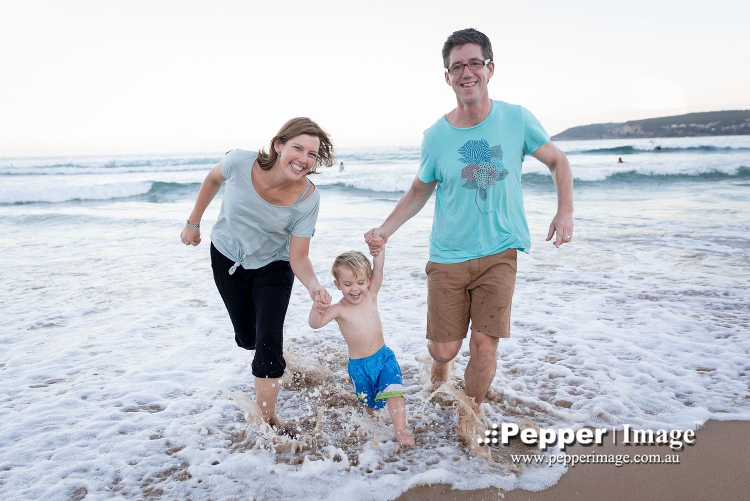 Happy family of three playing in ocean waves at the beach during sunset.