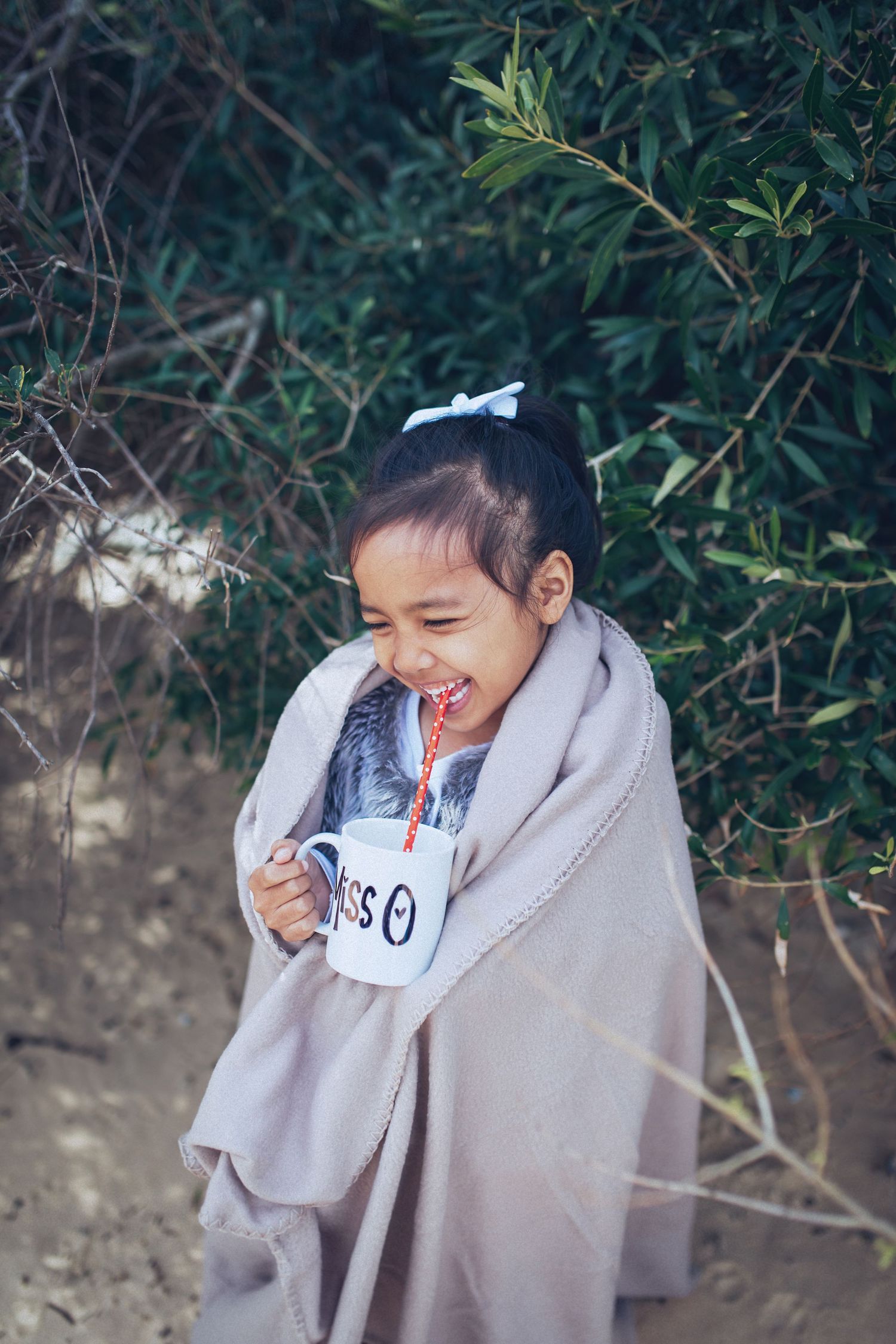 Laughing little girl wrapped in a beige blanket holding a white mug outdoors near green shrubs.