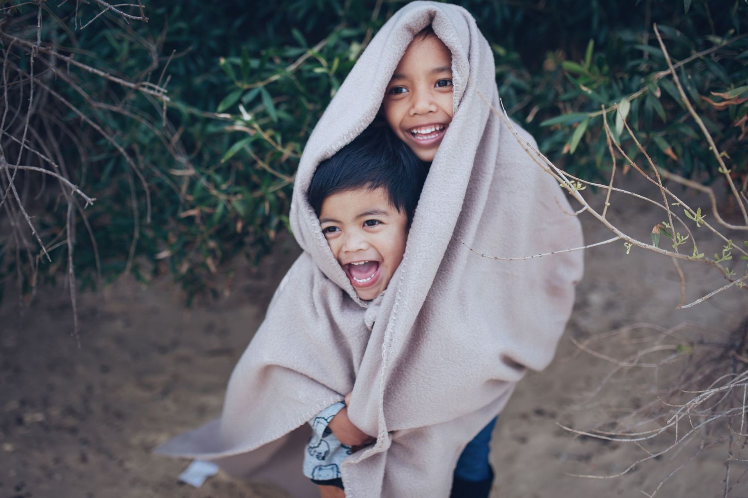 Two joyful children wrapped in a beige blanket laughing together in an outdoor natural setting.