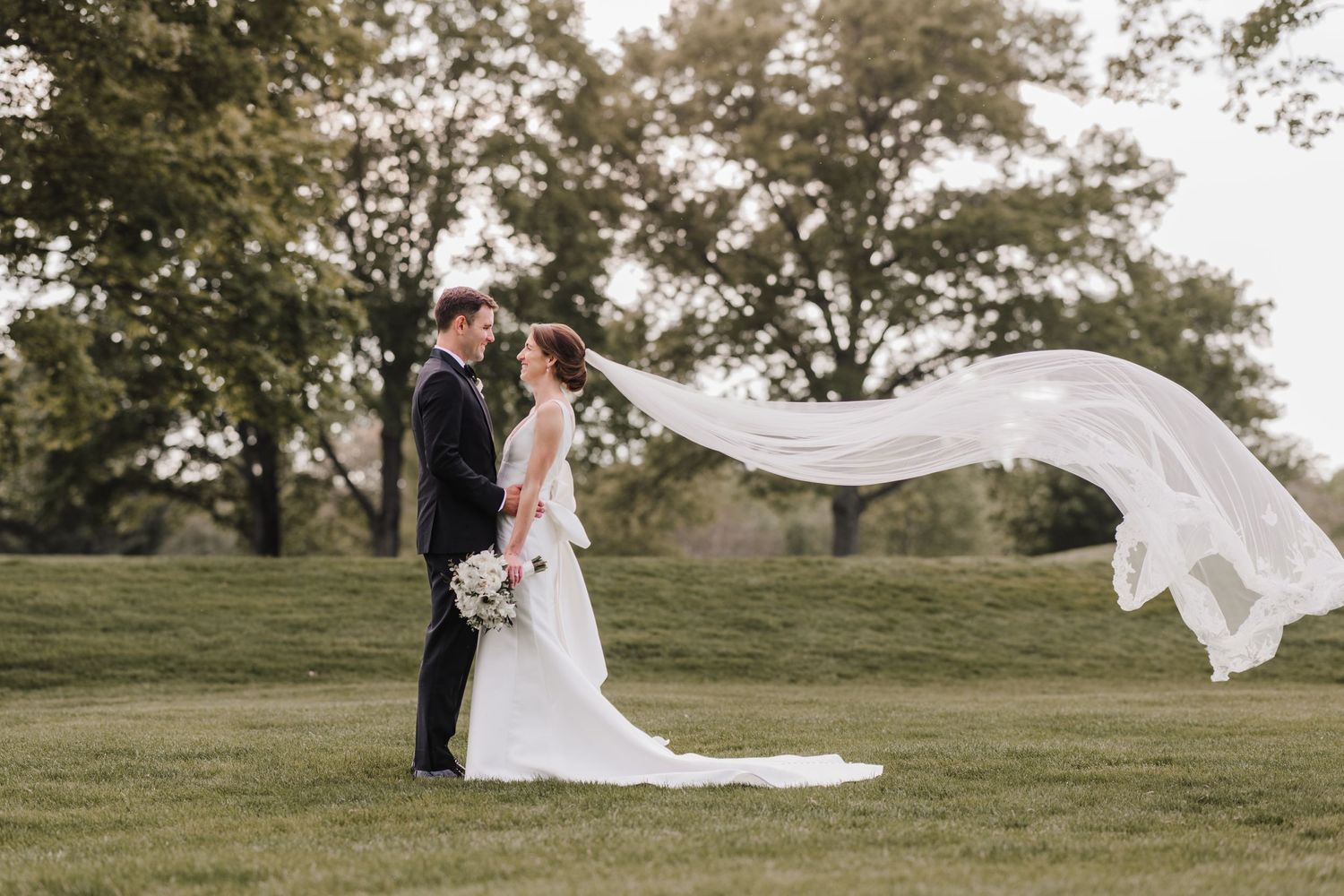 A romantic wedding photo taken outdoors with a flowing white veil catching the breeze on a green lawn.