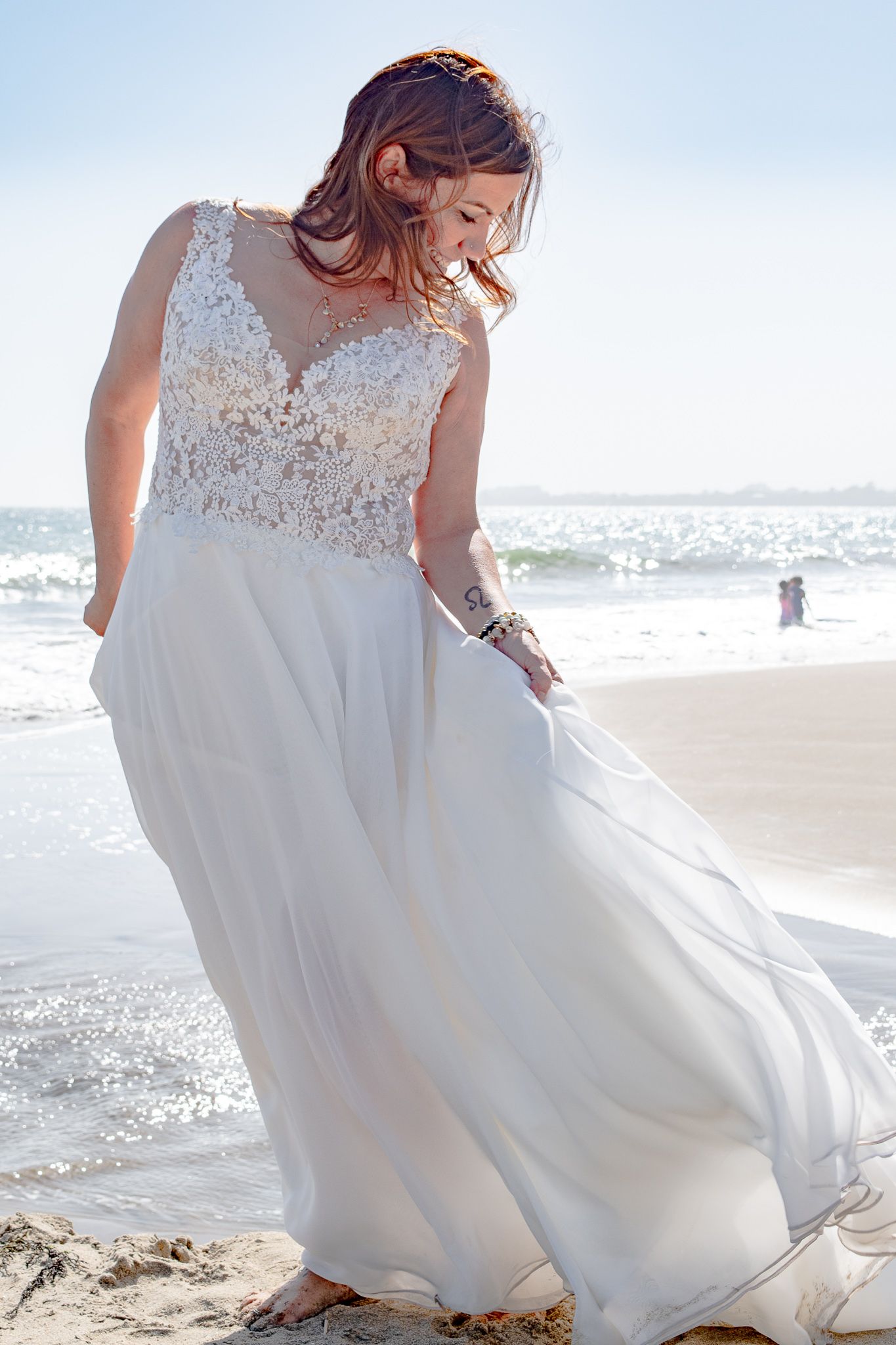Woman in wedding dress standing on the beach wind blowing hair, looking down at sand. Waves crashing behind her