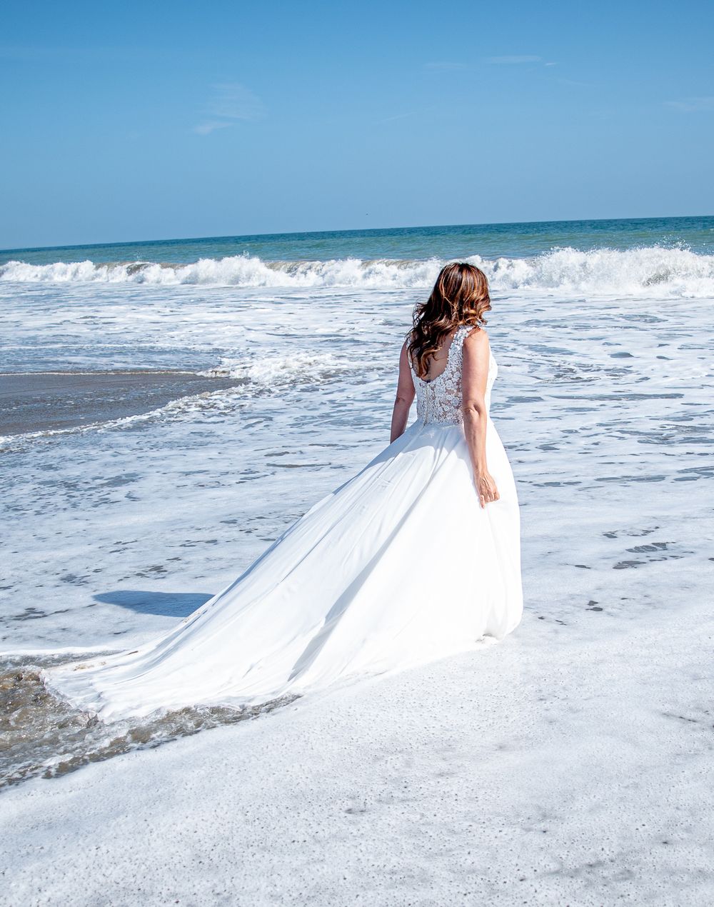 Woman in wedding attire walking out to sea. Waves carrying her dress. Waves crashing ahead.