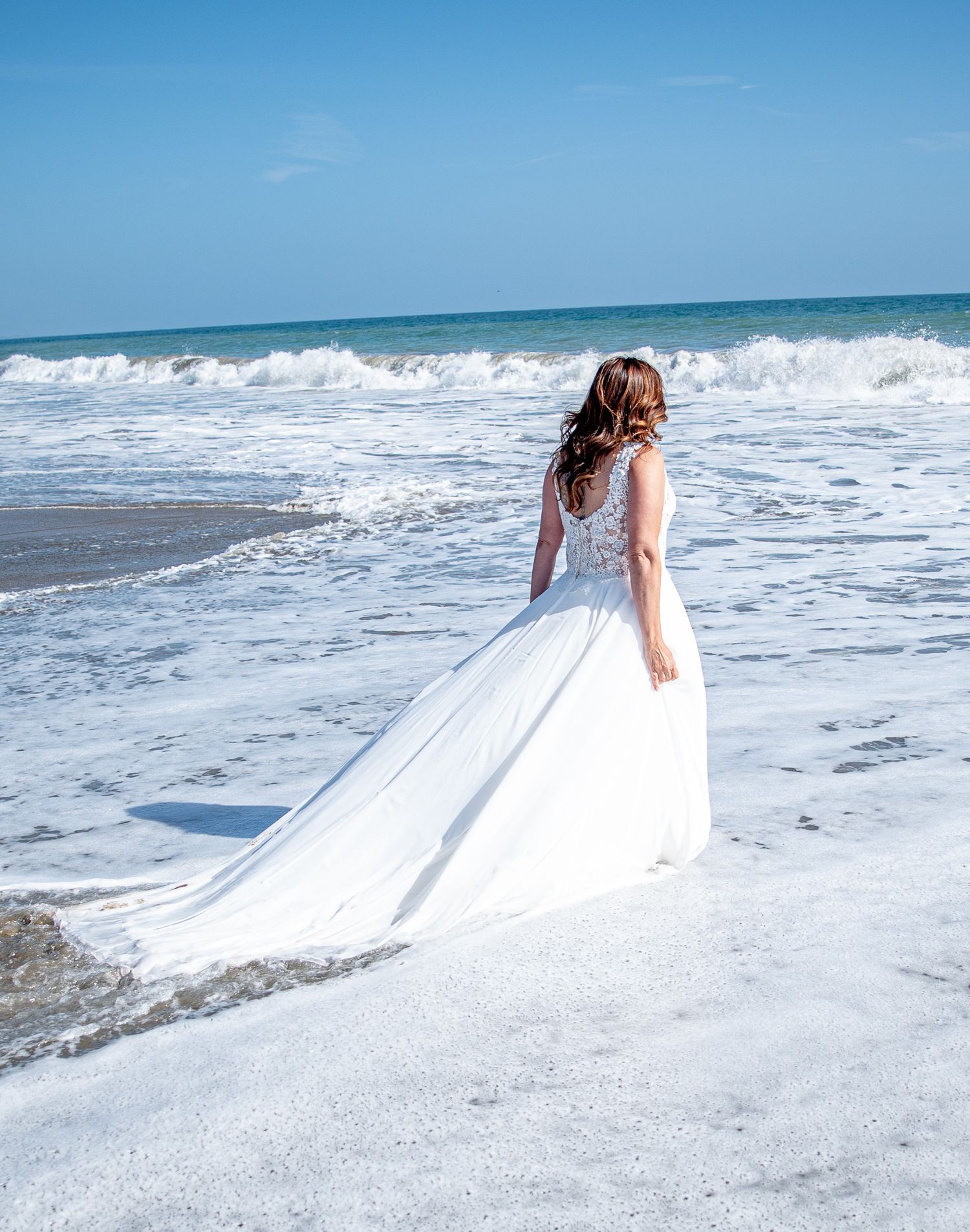 Woman in wedding attire walking out to sea. Waves carrying her dress. Waves crashing ahead.