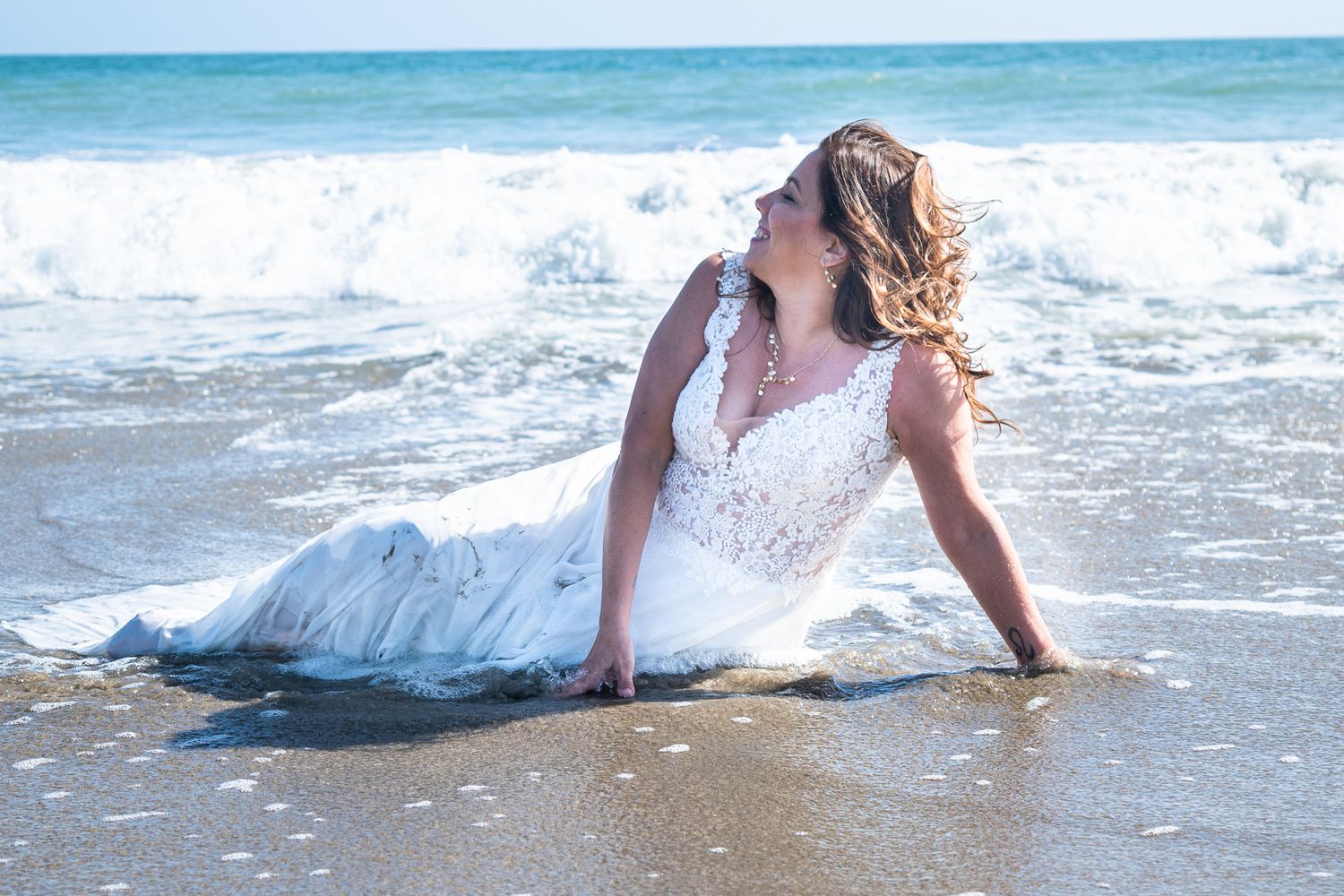 Woman in wedding dress sitting in the surf wind blowing hair, looking out to sea. Waves crashing behind her Mermaid pose
