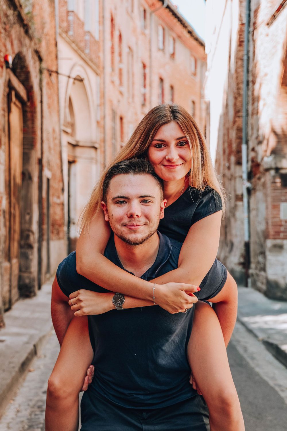 Couple sharing a piggyback ride moment in a narrow European street with historic architecture.