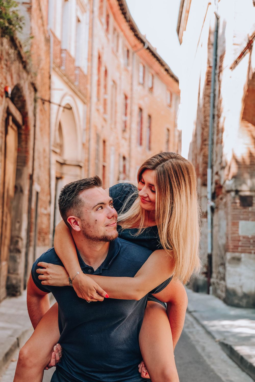 Couple enjoying a playful piggyback ride in a rustic European alley at sunset.