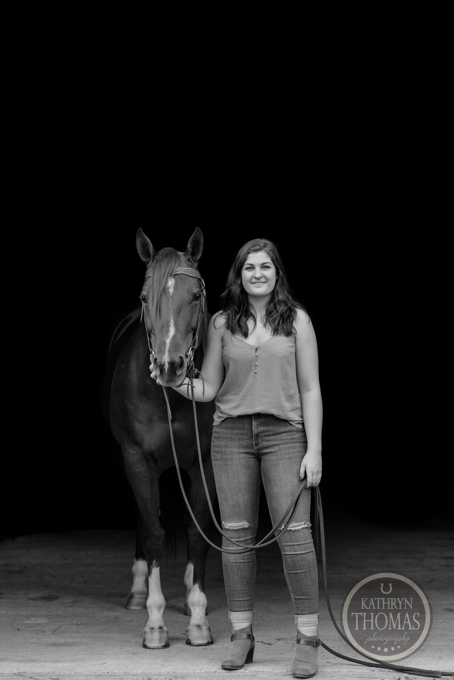 A black and white portrait of a person standing with their horse against a dark backdrop.