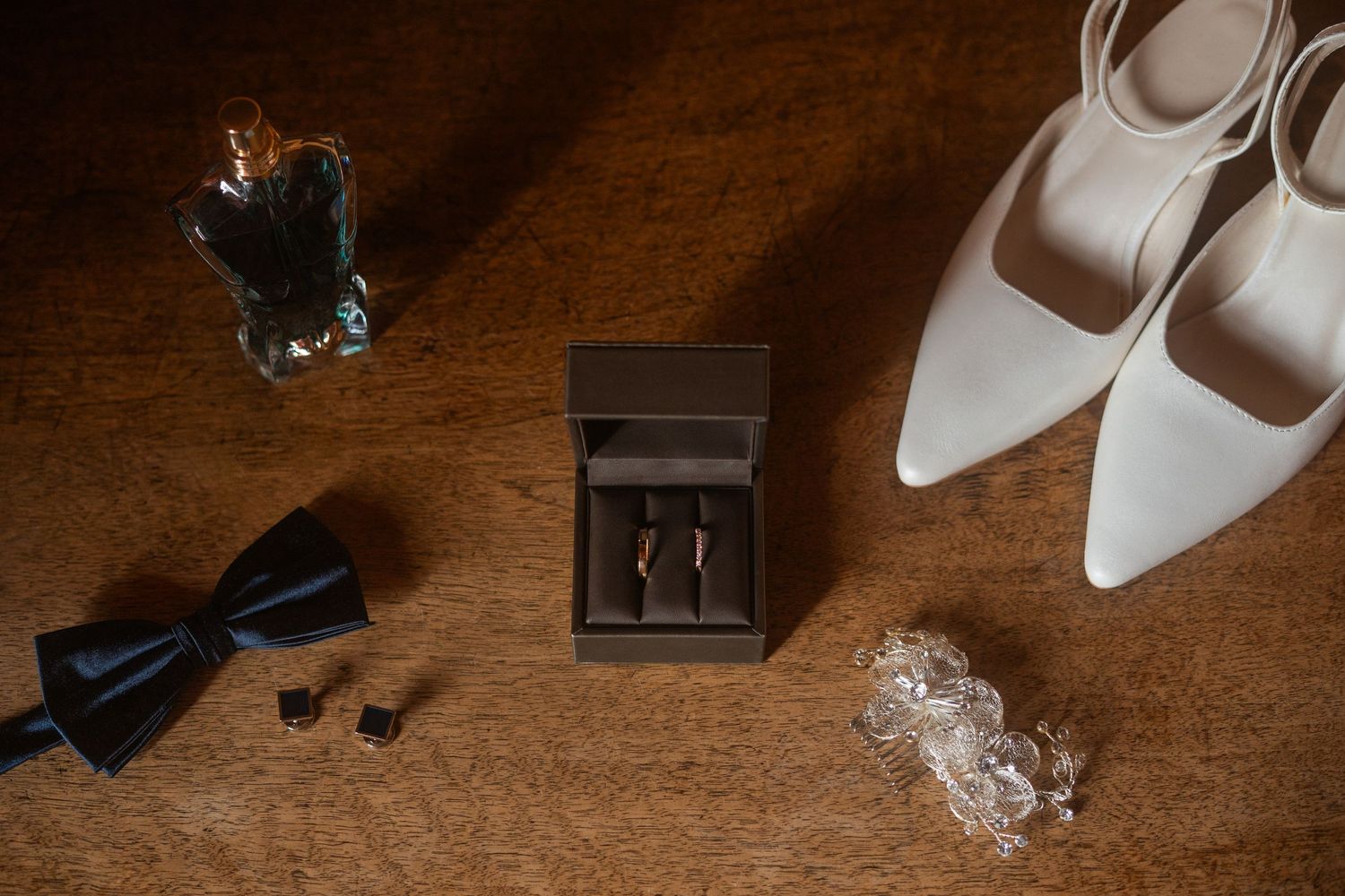 Wedding accessories including white shoes, bow tie, and rings arranged on rustic wooden surface.