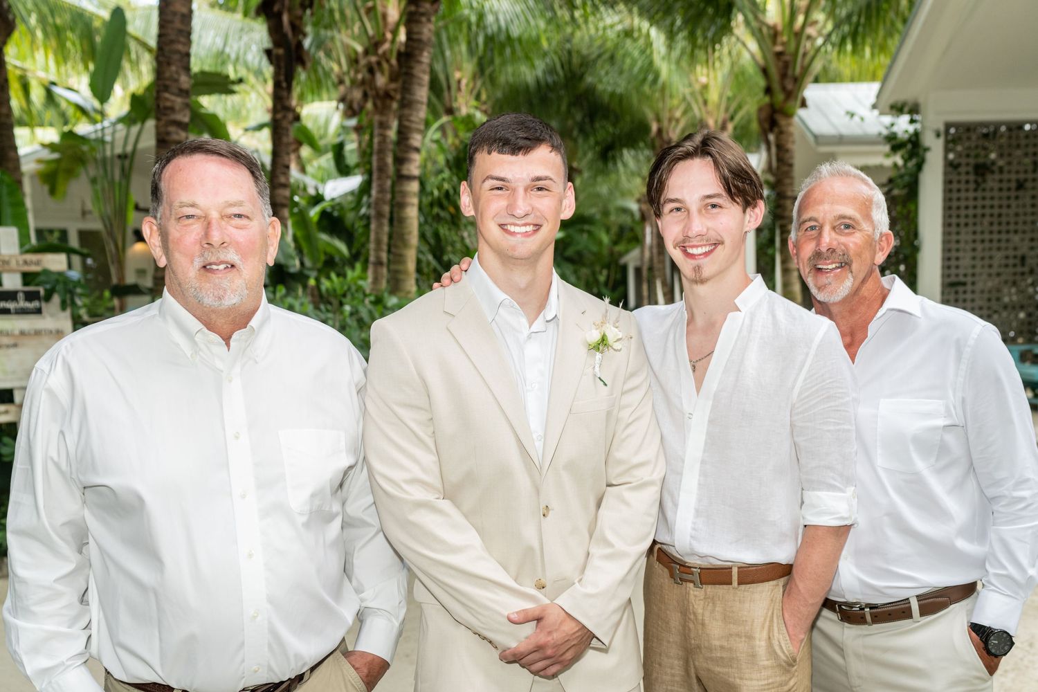 Four men in white and beige attire pose together outdoors at a tropical location.