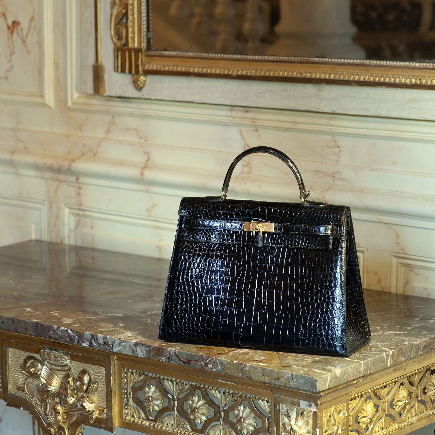 Sac à main de luxe (HERMÈS) en cuir de crocodile exposé sur une table en marbre doré orné dans une pièce élégante.