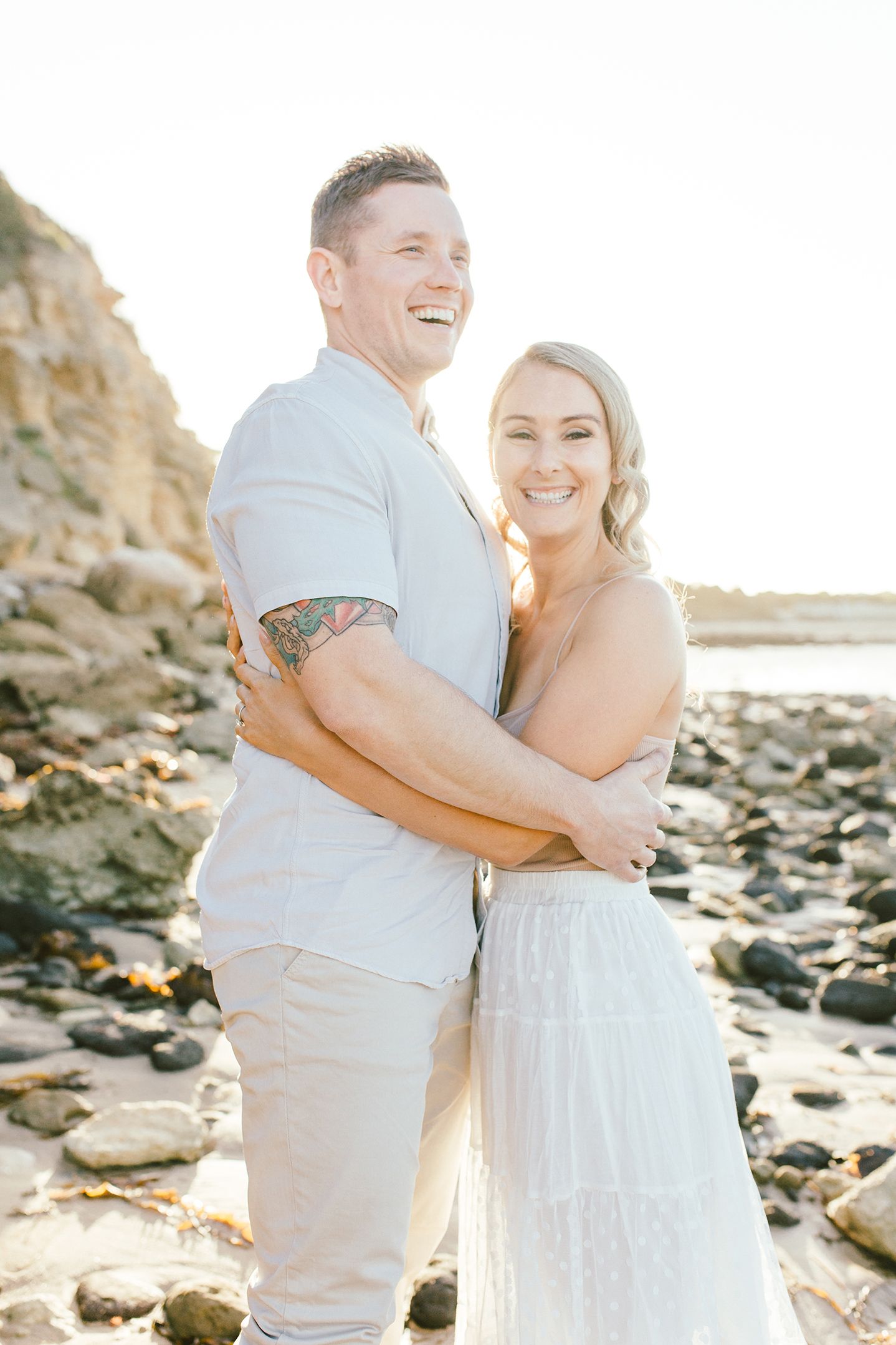 A romantic couple embraces and laughs together on a rocky beach during golden hour.