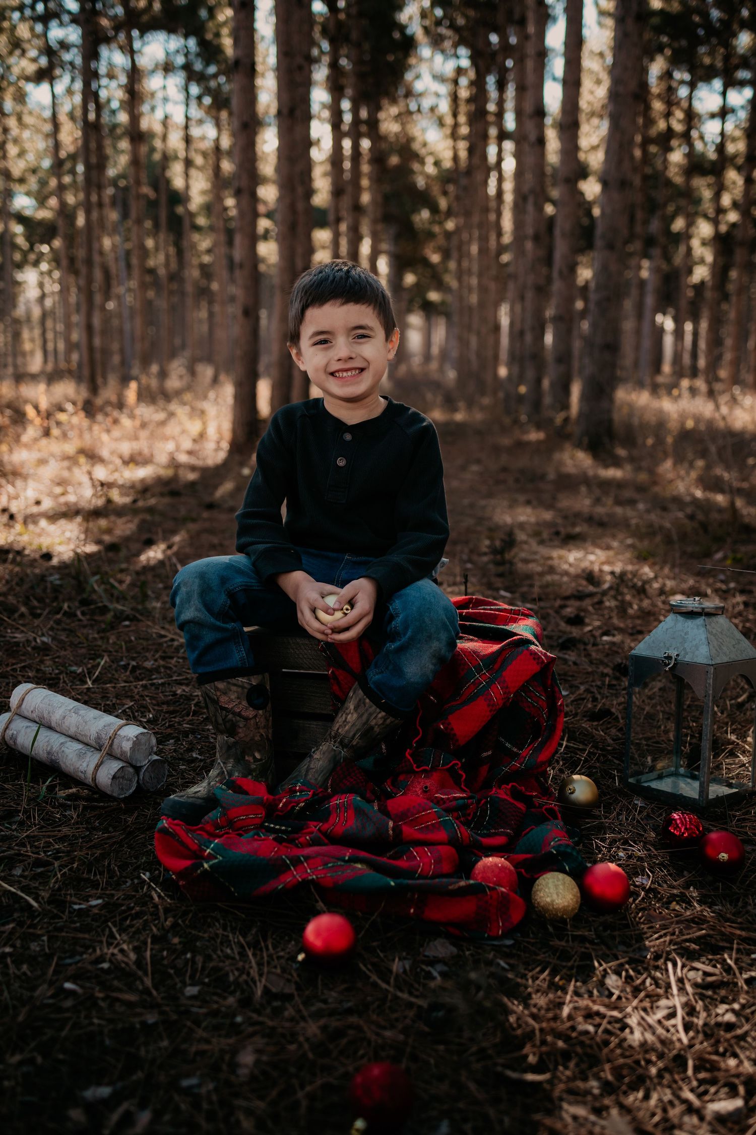 Young child sitting among holiday decorations and plaid blankets in a forest setting.