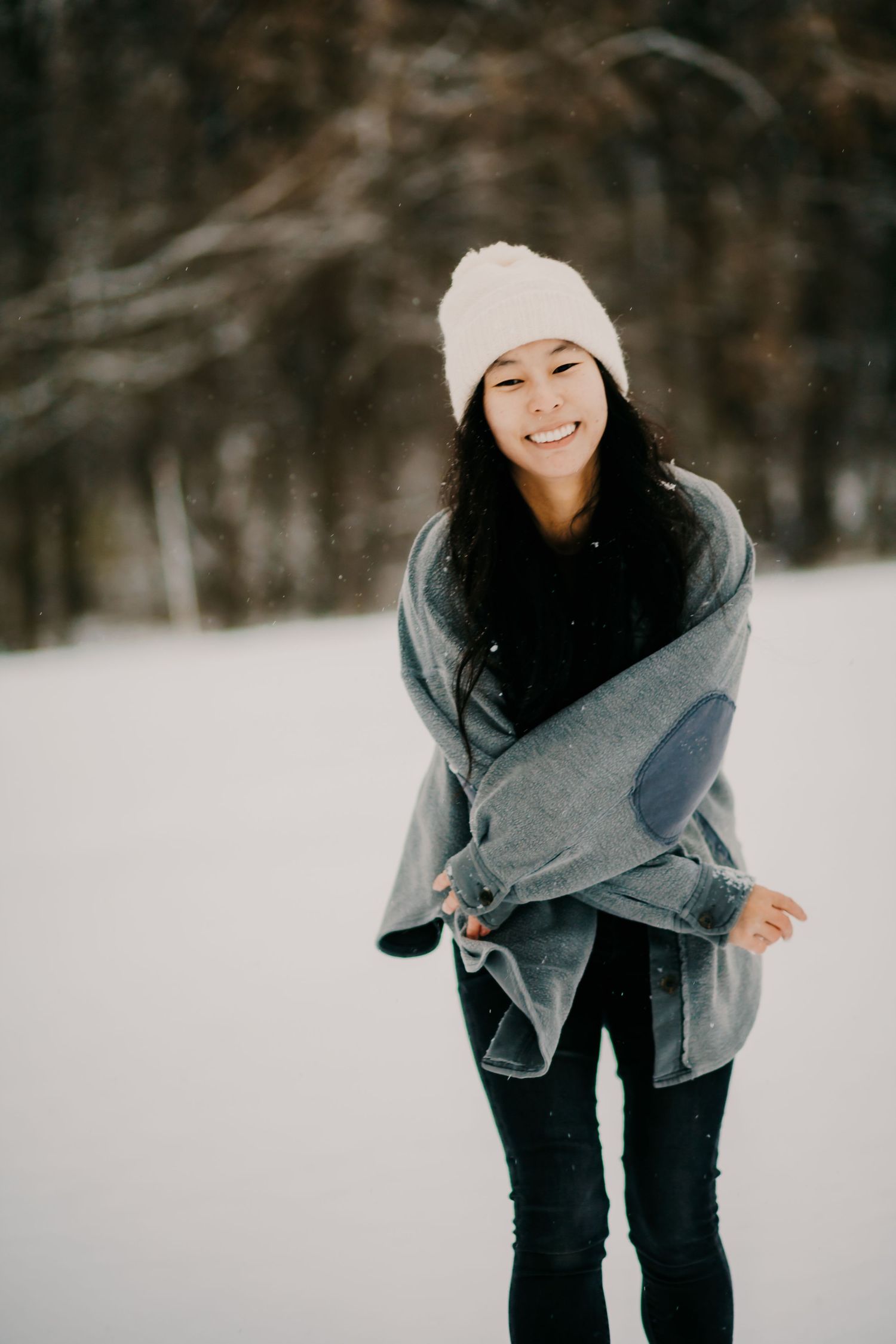 A person in winter attire smiles while wrapped in a gray blanket in a snowy setting.