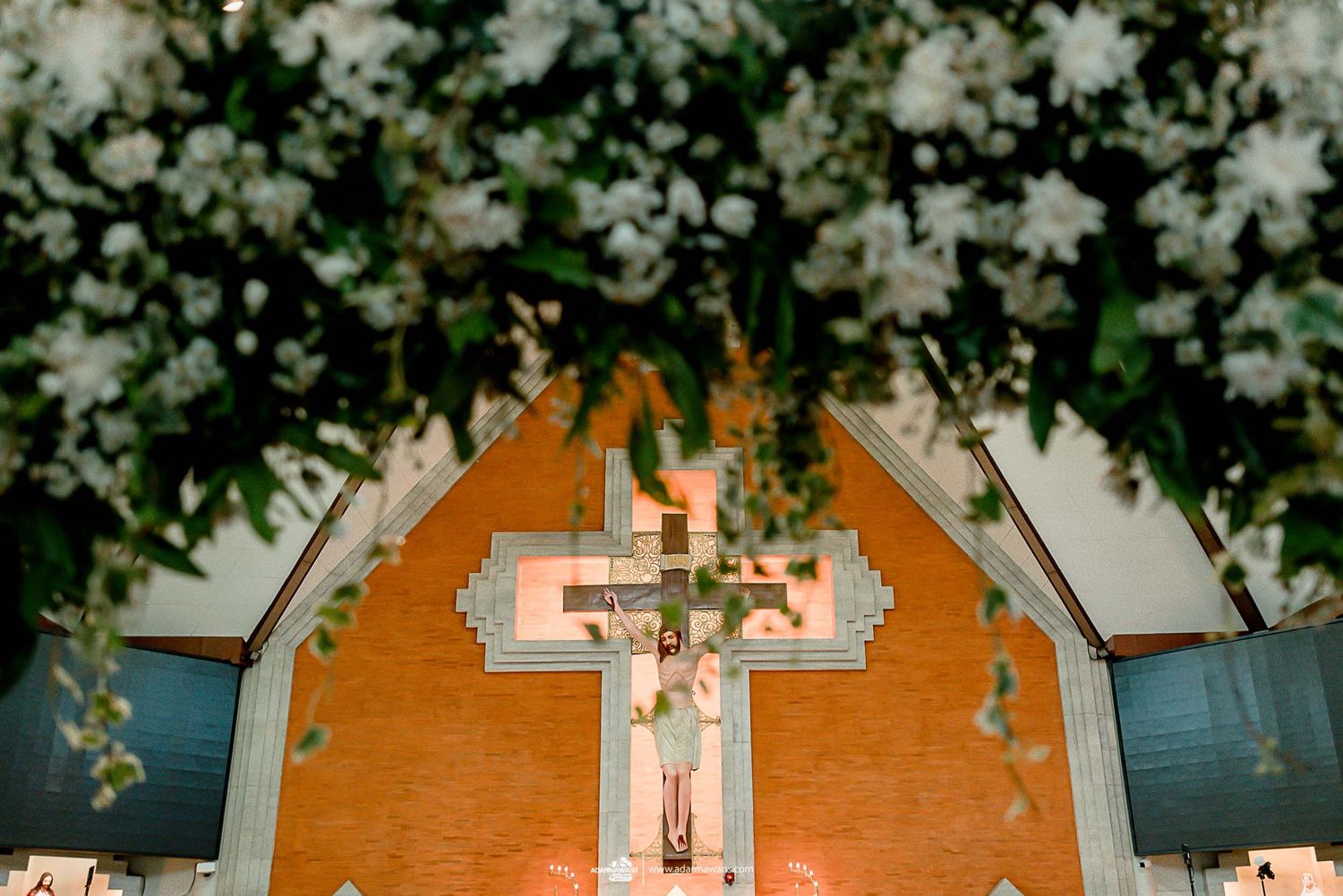 Church interior with white flowers and illuminated crucifix against orange backdrop.