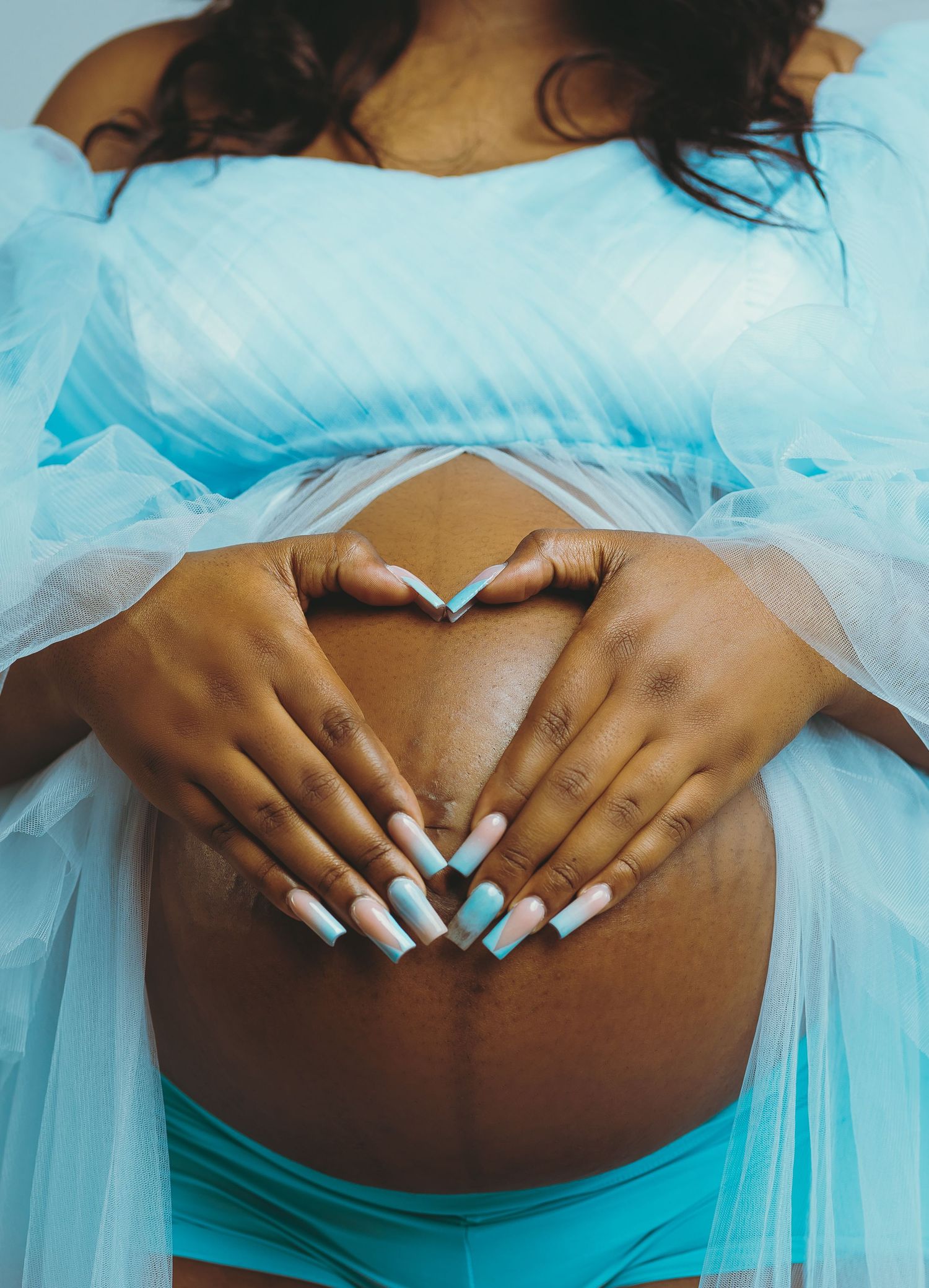 Maternity photo showing hands forming heart shape over pregnant belly with blue tulle fabric.