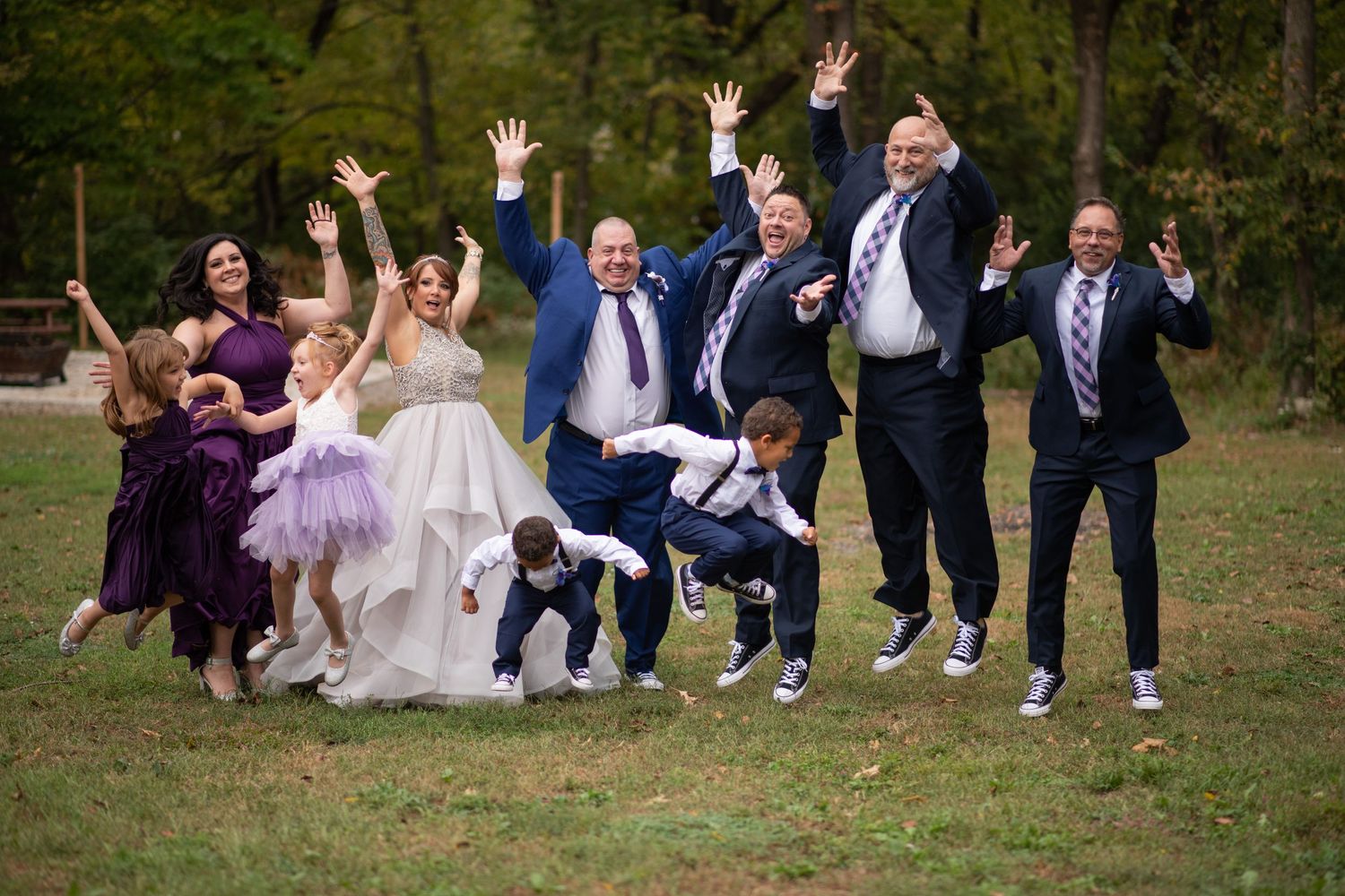 Wedding party jumps joyfully for a fun group photo on grass.