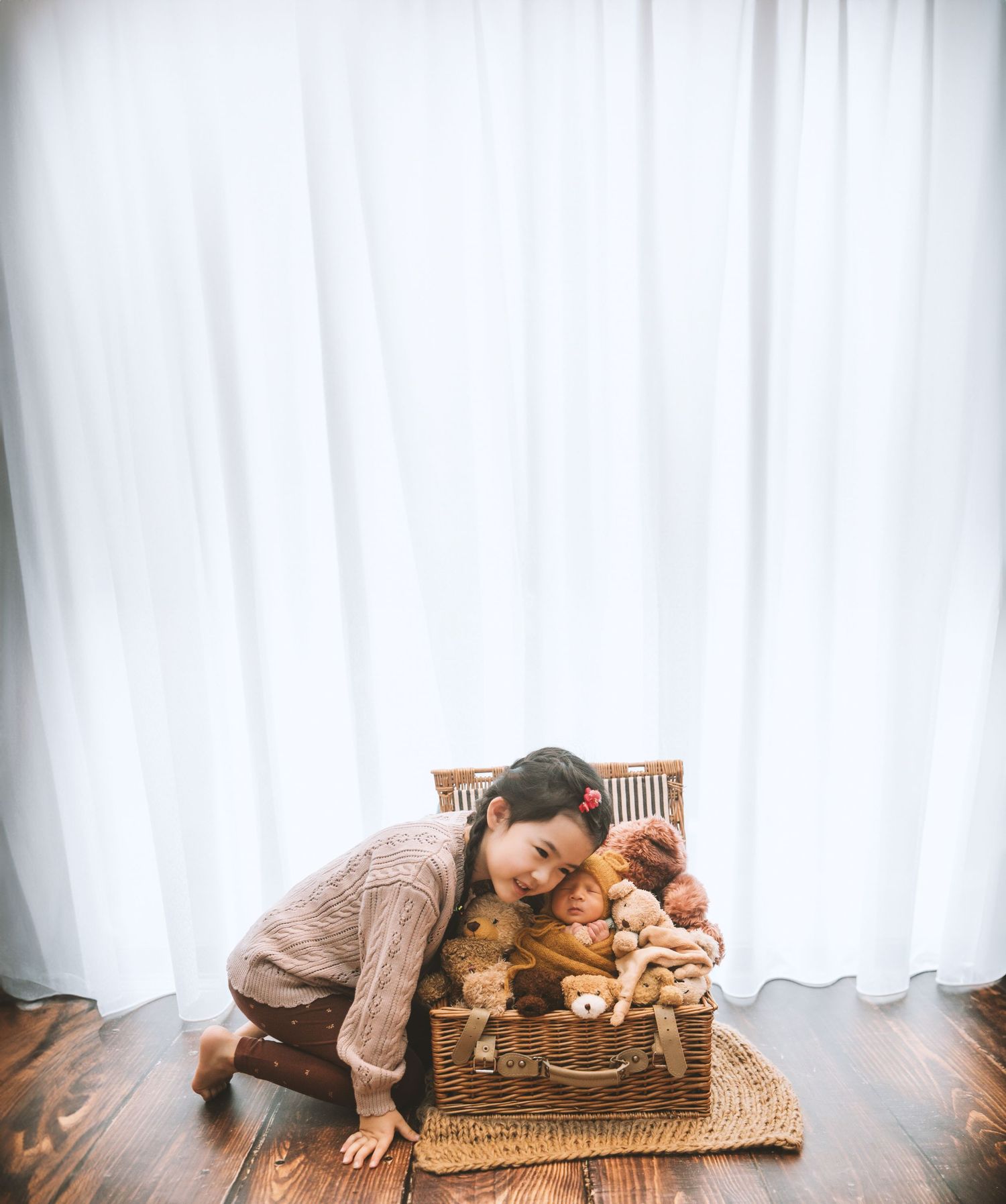 Smiling mother kneeling beside large wicker basket filled with sleeping newborn twins and stuffed bears.