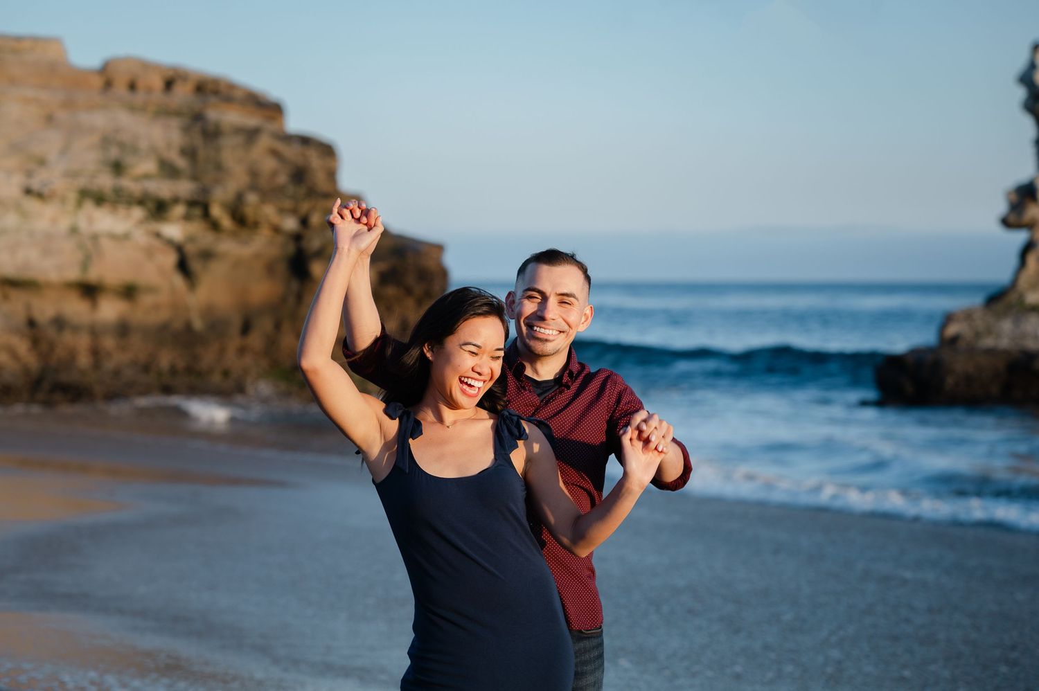 A joyful couple dances together on a beach with coastal rock formations behind them.