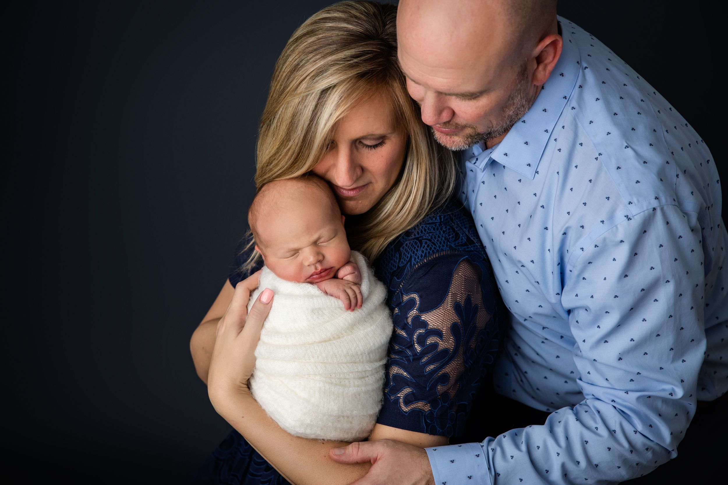 Studio portrait of couple tenderly holding swaddled newborn against dark background.