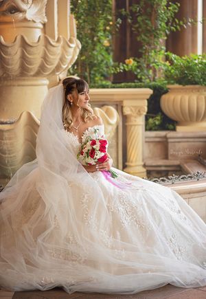 Bride in white lace gown holding red bouquet poses by ornate stone pillars.