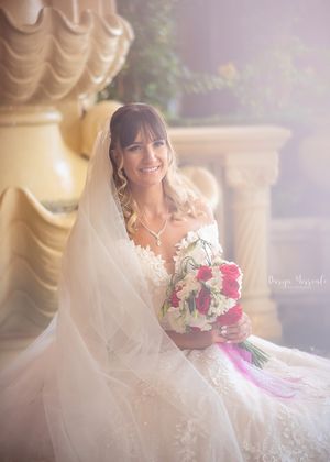 A bride in a lace wedding dress and veil holds a pink and white bouquet while sitting in soft lighting.