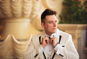 A groom in a white tuxedo adjusts his bowtie while posing near classical columns.