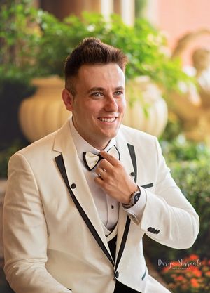 A groom in a white and black tuxedo smiles while adjusting his tie outdoors.