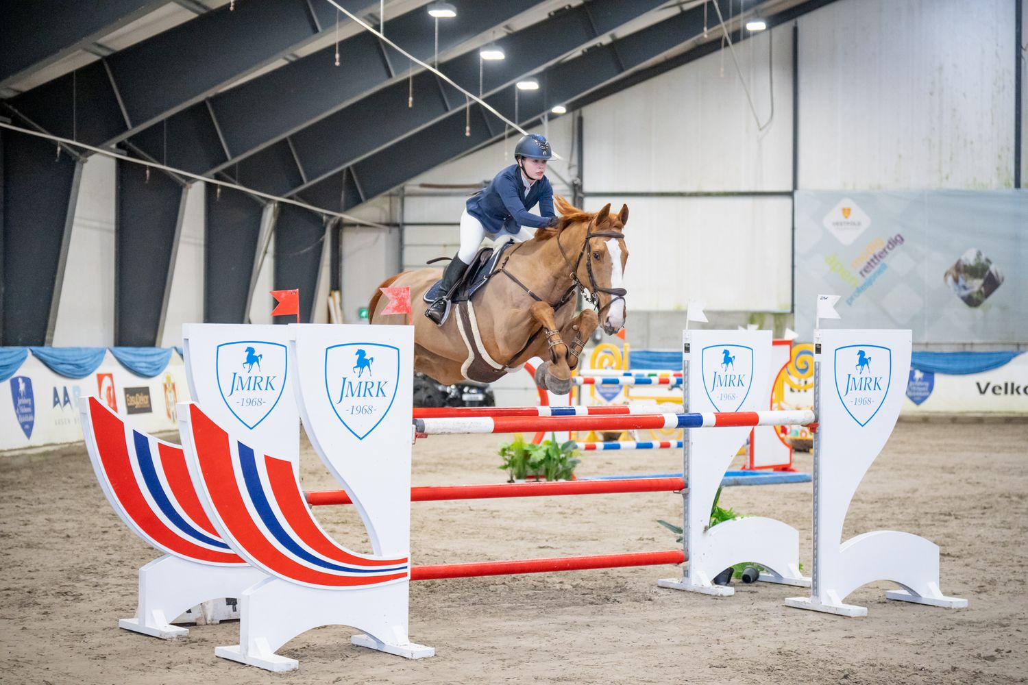 Horse and rider jumping over colorful red and white obstacles in an indoor arena.