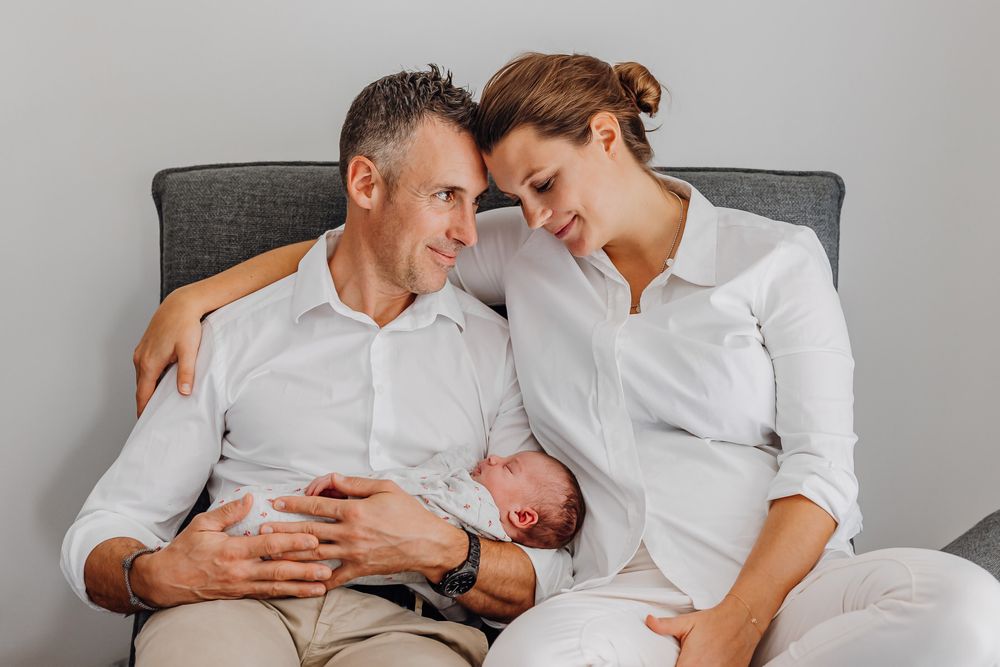 Zarter Familienmoment auf grauer Couch in passenden weißen Outfits.