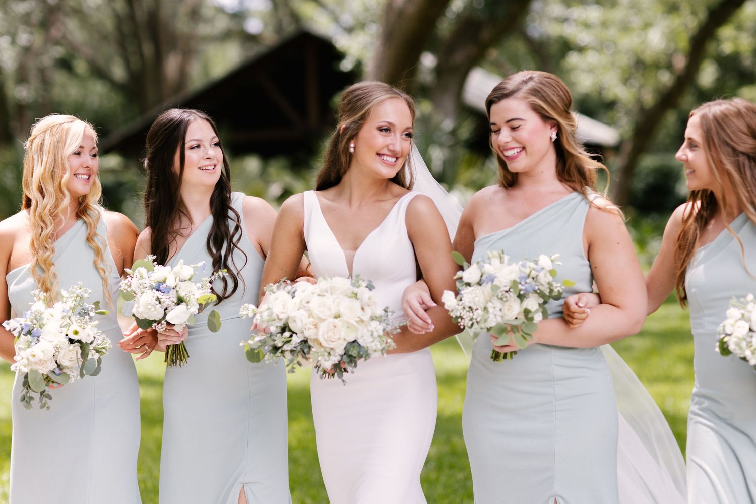 Bridesmaids in light blue dresses holding white bouquets share laughs during wedding celebration.