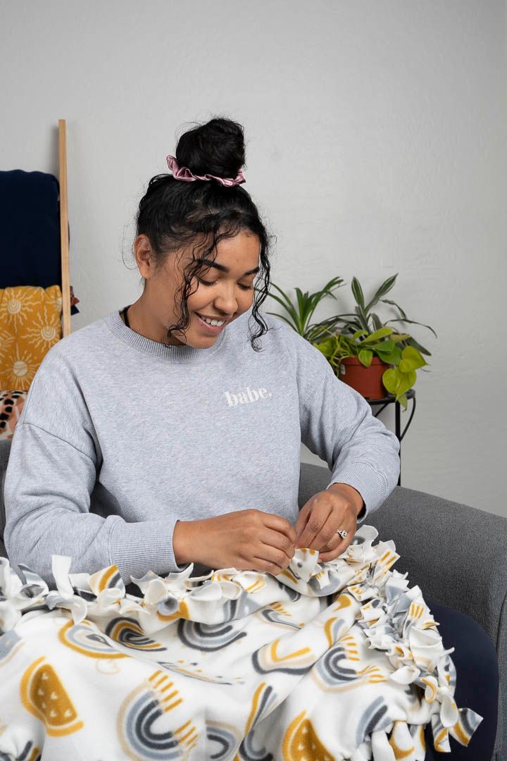 Someone works on a handmade fleece blanket project while wearing a gray sweatshirt.