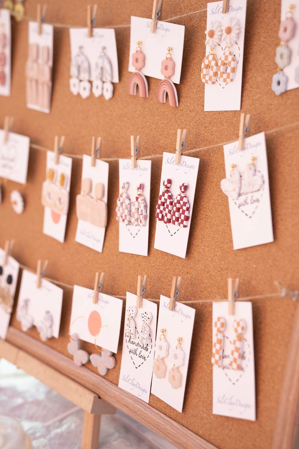 Handmade earring designs displayed on white cards hanging from twine against a cork board.