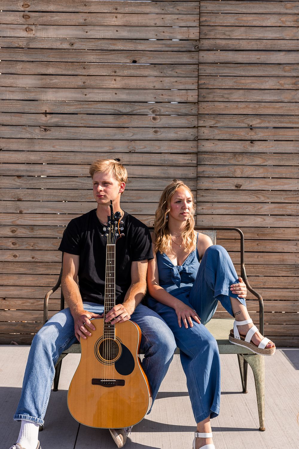 Musicians with acoustic guitar sitting on bench against wooden wall in casual attire.